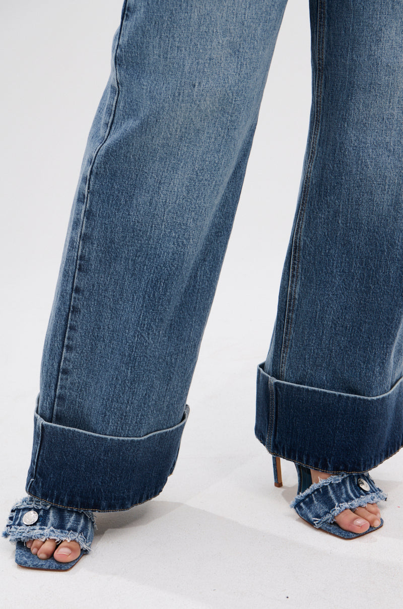 Close-up of someone in JORDAN WIDE LEG DENIM—high-rise blue jeans with a wide-leg, cuffed hem—paired with matching denim heeled sandals that show bare toes and feet, set against a simple light background.
