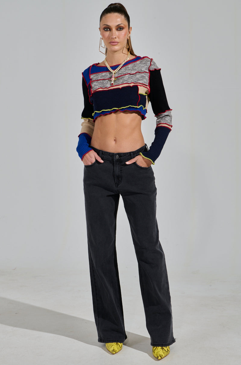 A person with slicked-back hair poses confidently, hands in pockets, wearing a colorful cropped sweater and NICOLE VINTAGE LOW RISE JEAN. Yellow snakeskin-patterned shoes add a bold touch against a plain, minimalistic backdrop.