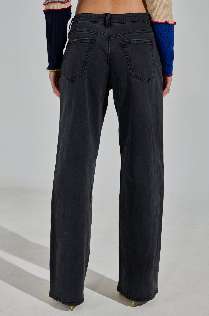 Dressed in NICOLE VINTAGE LOW RISE JEAN and a long-sleeve color-block sweater, a person reveals the detailed back pocket design. The background remains plain and minimalistic.