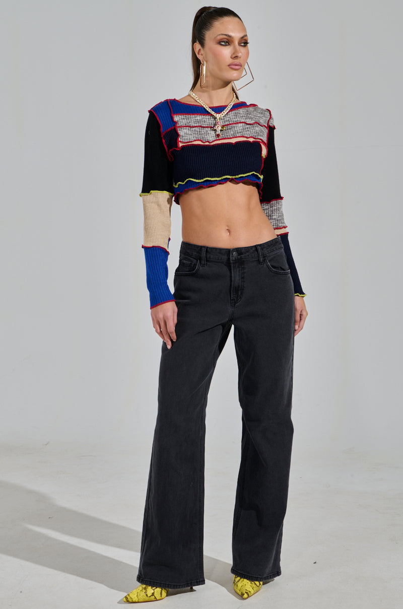 A person stands confidently wearing a multicolored long-sleeve crop top and the NICOLE VINTAGE LOW RISE JEAN in black. With a ponytail and yellow pointed shoes, they pose against a plain, light gray wall.