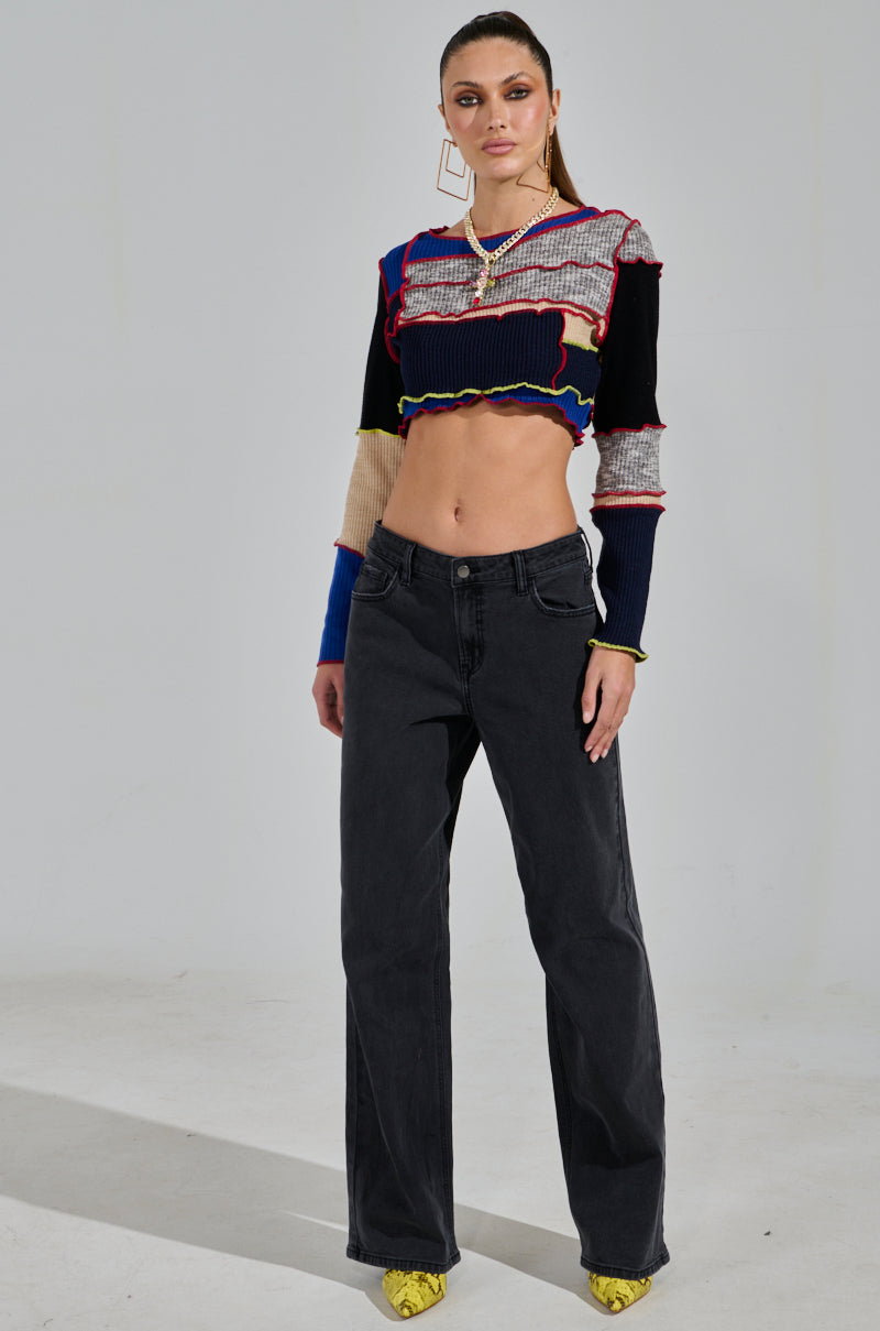 A woman poses confidently against a plain backdrop, showcasing a colorful patchwork crop top, NICOLE VINTAGE LOW RISE JEAN, statement earrings, and yellow shoes. Her hands rest relaxed at her sides with her hair styled back.