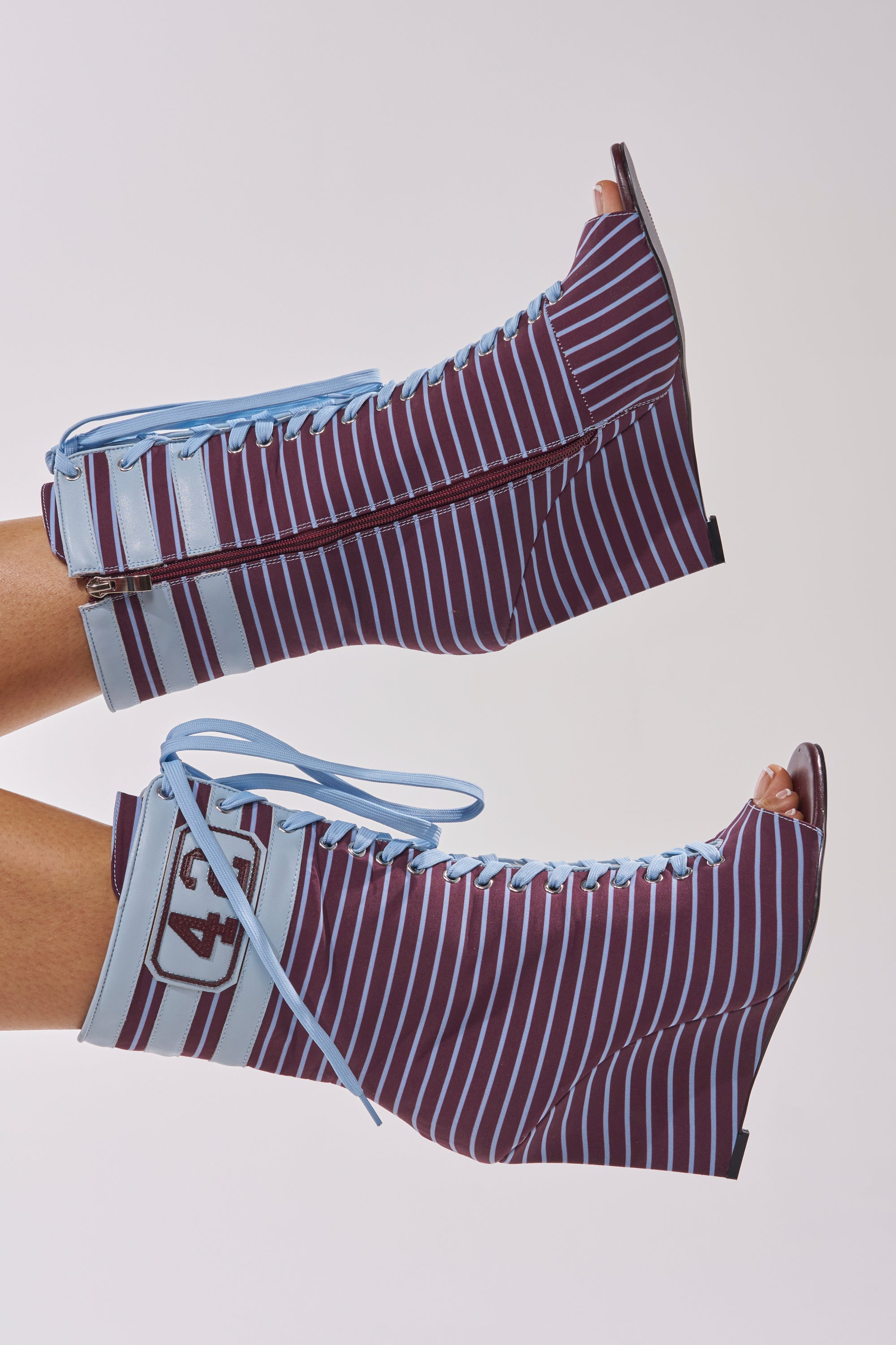 Two feet in AZALEA WANG WYATT BLUE MULTI STRIPE WEDGE BOOTIES with maroon and light blue vertical stripes, lace-up fronts, side zippers, and a 43 patch, shown on a light background—a bold courtside streetwear look.