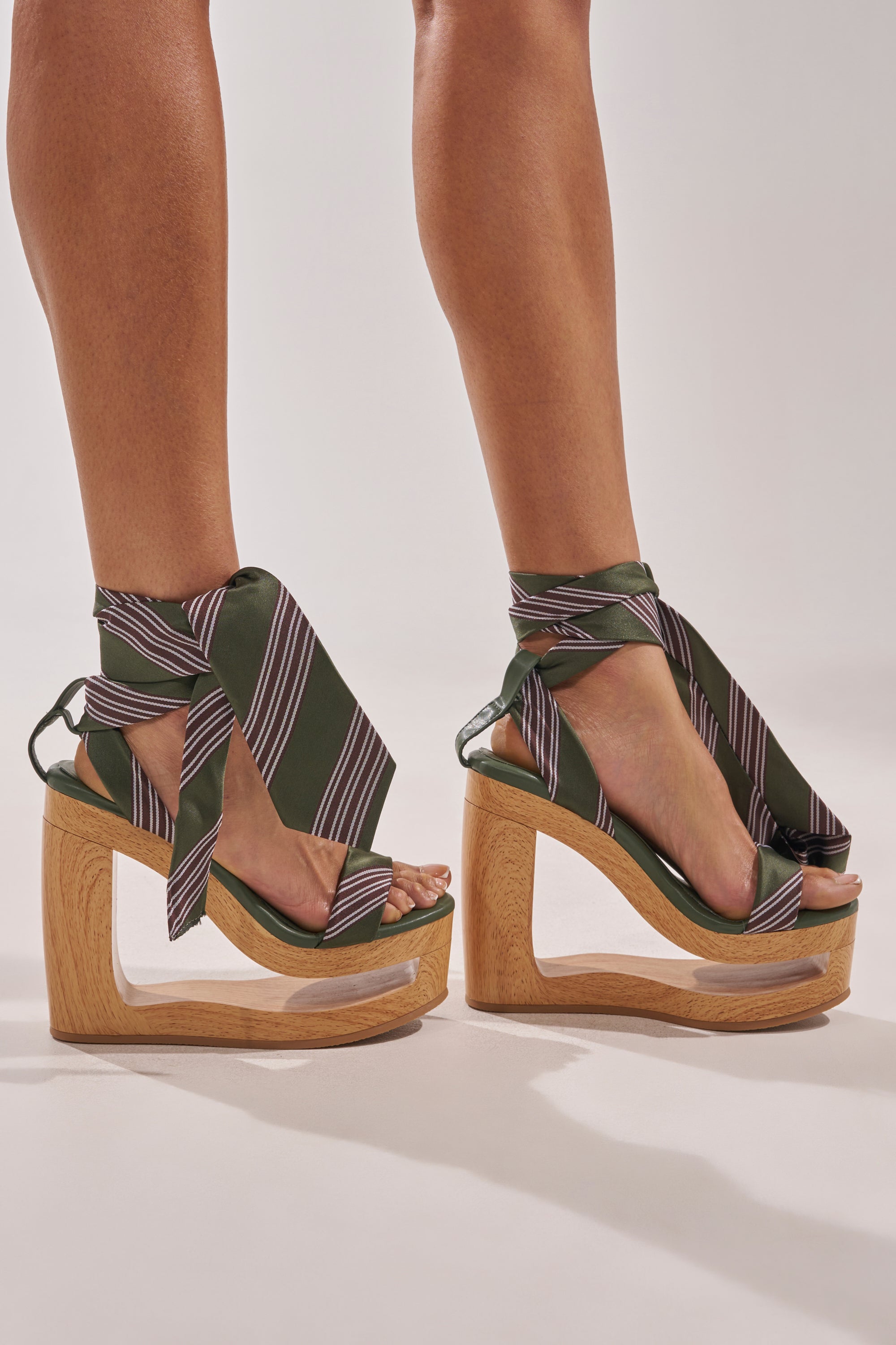 Wearing the AKIRA Gold Wynn Green Tie Lace Up Novelty Wedge Sandal with chunky wooden platform soles and green ankle-wrapping straps, a person stands against a plain light background. 0