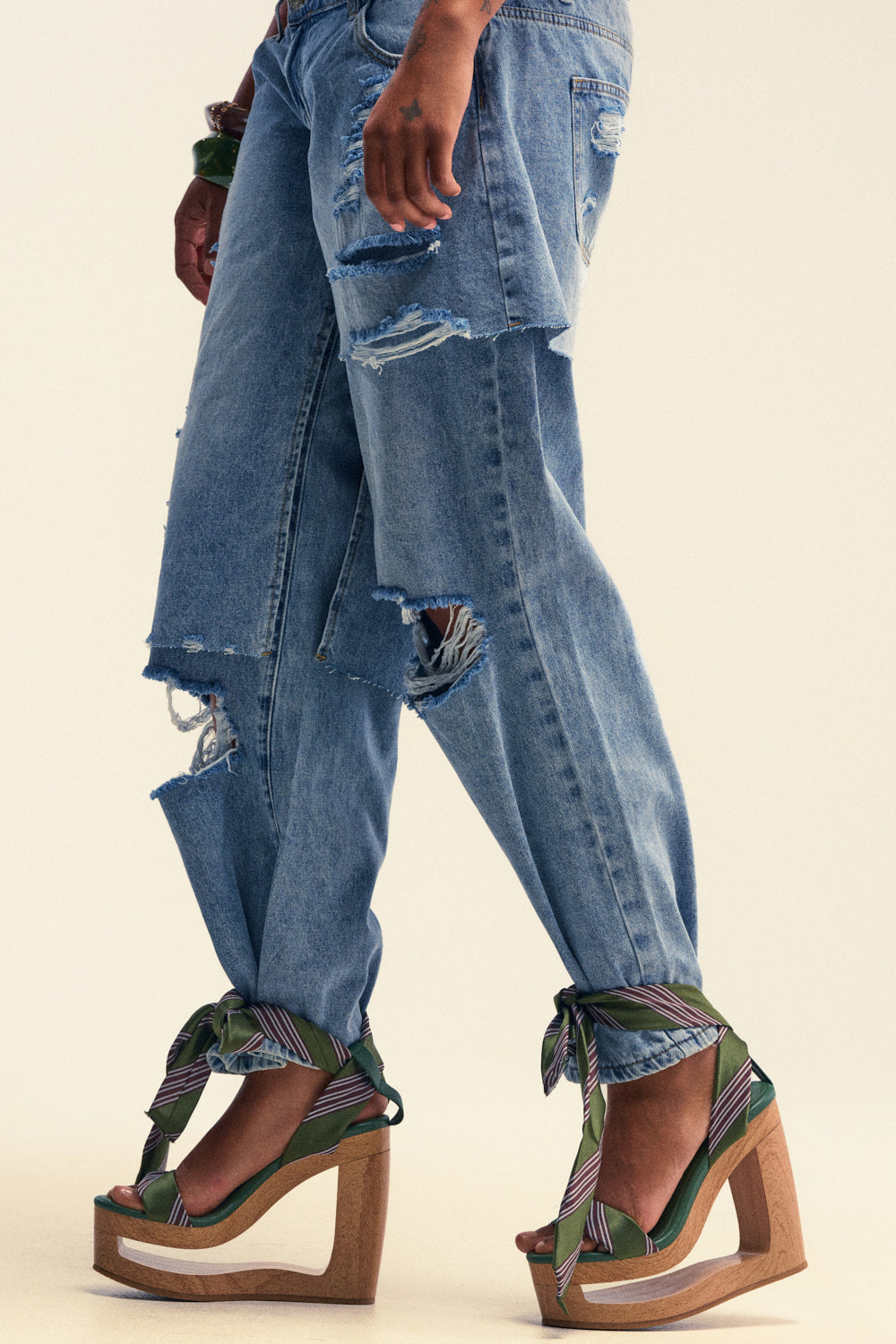 A person wearing ripped blue jeans and the AKIRA GOLD WYNN GREEN TIE LACE UP NOVELTY WEDGE SANDAL, featuring green straps with a subtle bow detail, stands against a plain background.
