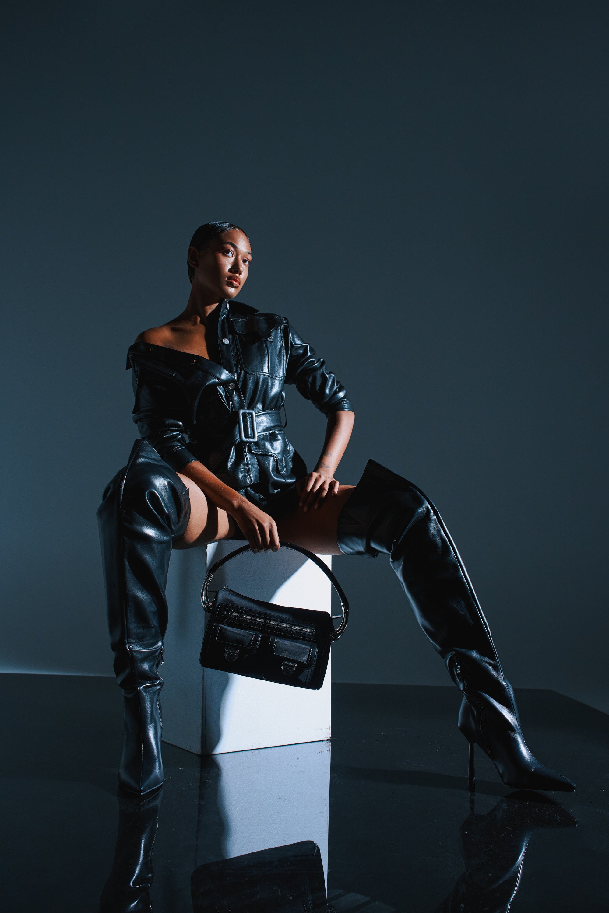 A woman poses confidently on a white block, wearing thigh high stiletto boots—the AZALEA WANG WYNNDY BLACK THIGH HIGH BOOT—styled with a belted faux leather jacket and black handbag. The dark background enhances the high-fashion vibe.