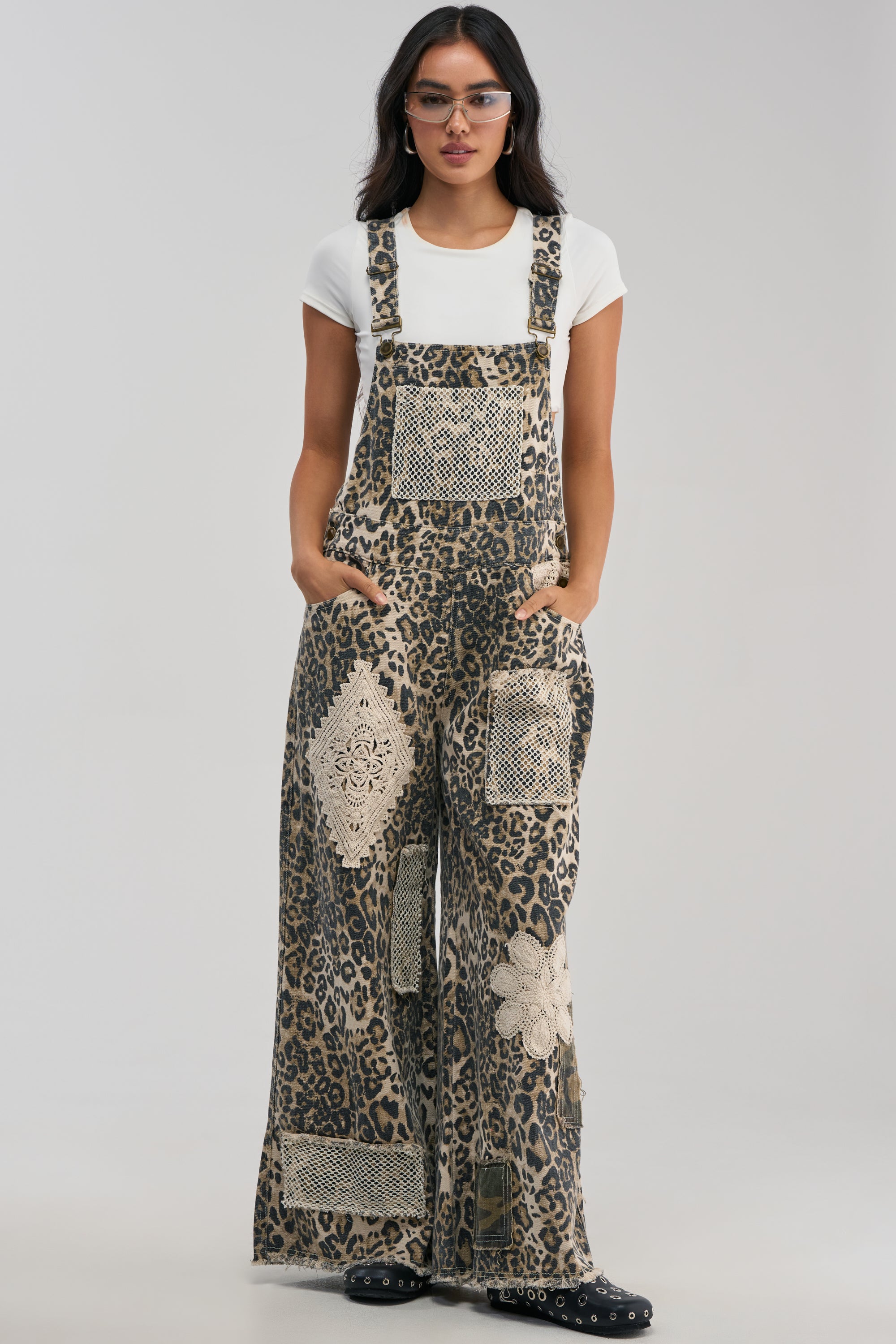 Wearing eyeglasses and a white t-shirt, this individual models the CATCH ME IN THE CITY LEOPARD OVERALLS featuring crochet patchwork, standing with hands in pockets against a plain light background—a standout choice for festival streetwear.