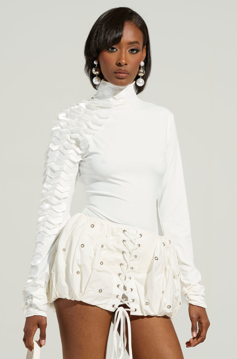 A woman in a chic white outfit showcases the NADIA Organza Long Sleeve Scale Top, characterized by geometric designs and ruffled accents on one side, paired with lace-up shorts. Her look is complemented by large pearl earrings and sleek, shoulder-length hair as she poses against a plain background.