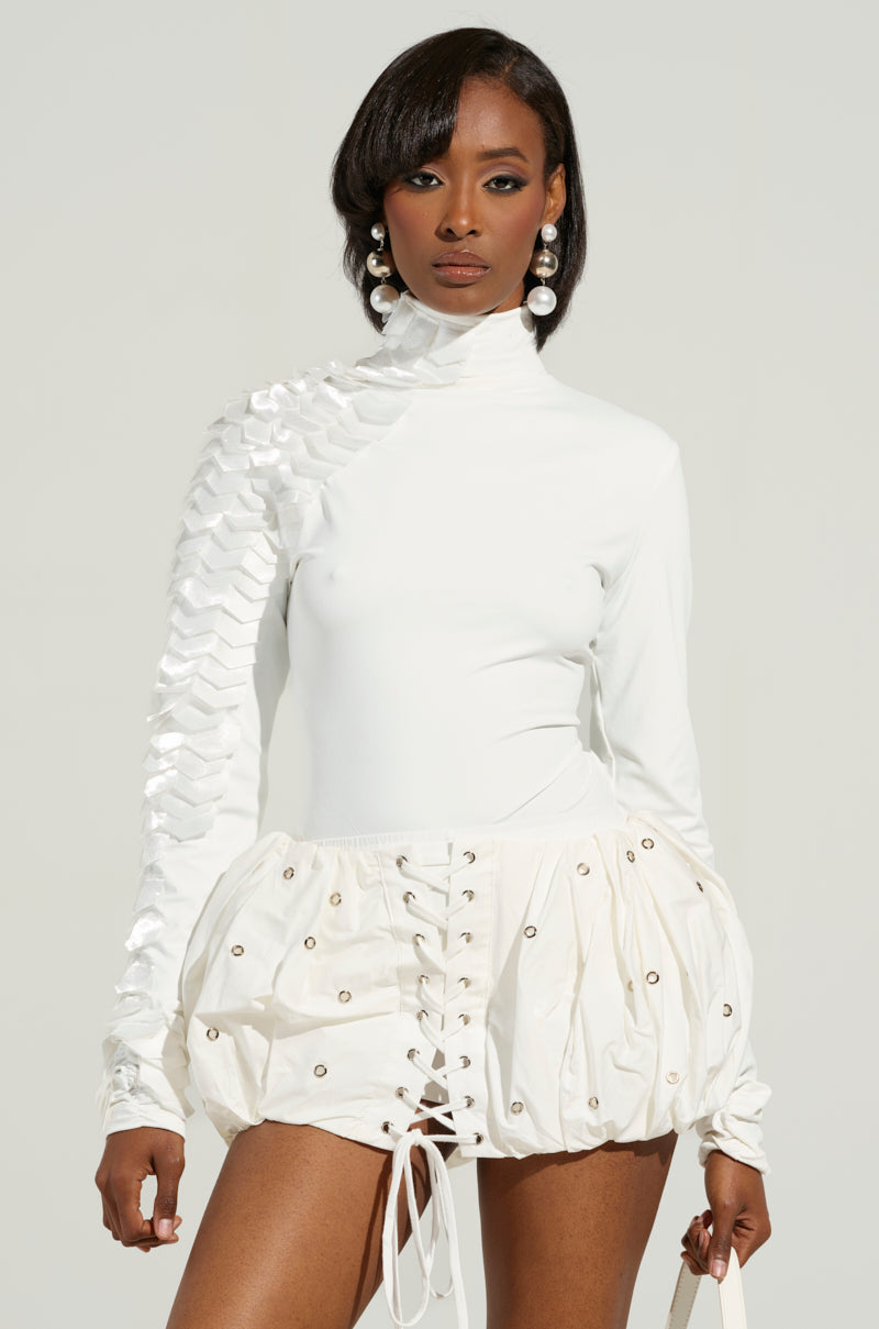 In a stylish white ensemble, a woman poses wearing the Nadia Organza Long Sleeve Scale Top, showcasing textured detailing on one shoulder. She pairs it with a mini skirt featuring a lace-up design and eyelets. Her look is complemented by large round earrings and a matching white bag, set against a neutral background.
