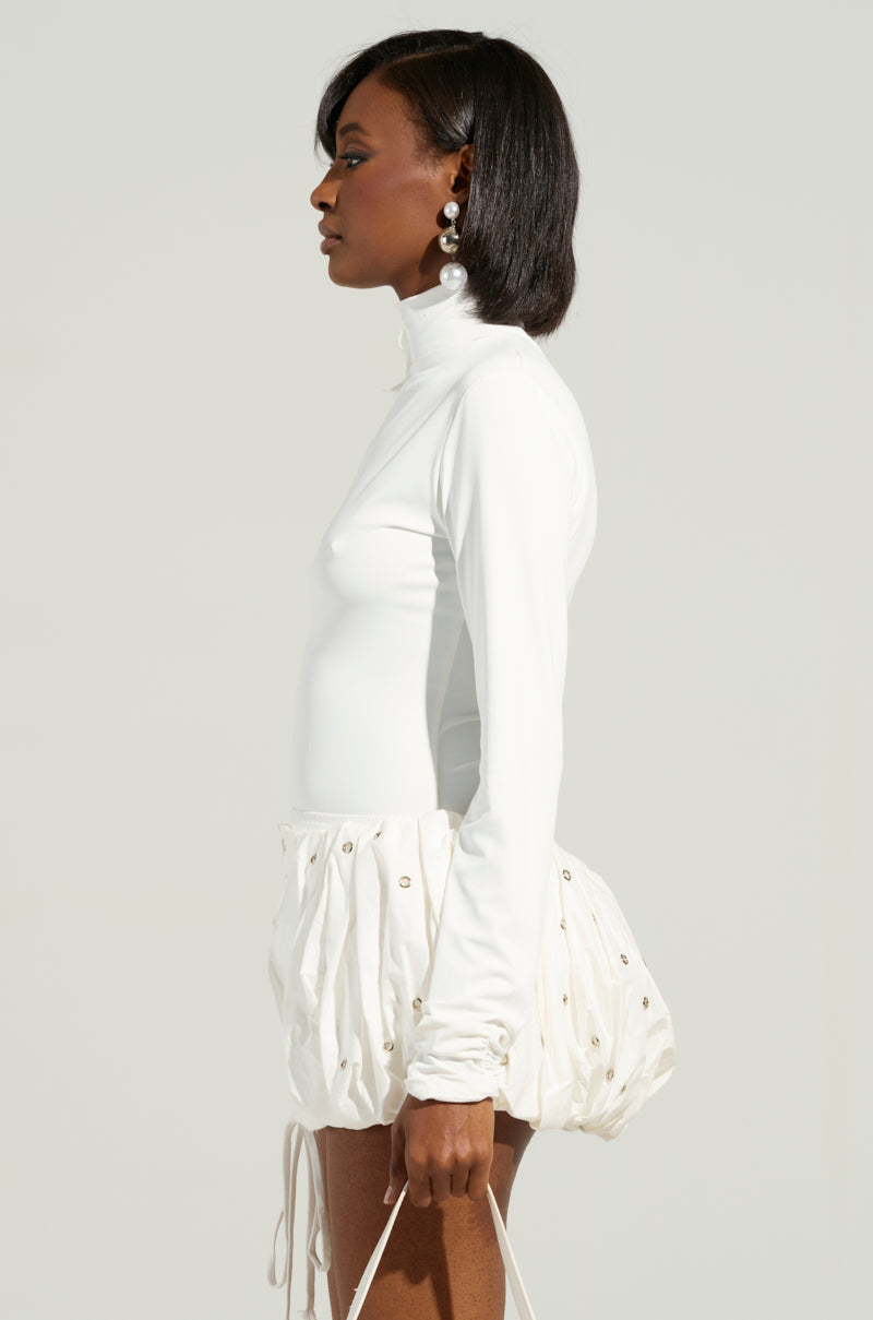 A woman with short hair stands in profile, dressed in the NADIA ORGANZA LONG SLEEVE SCALE TOP and a textured white skirt adorned with decorative elements. Her pearl earrings perfectly complement her style against a neutral background.