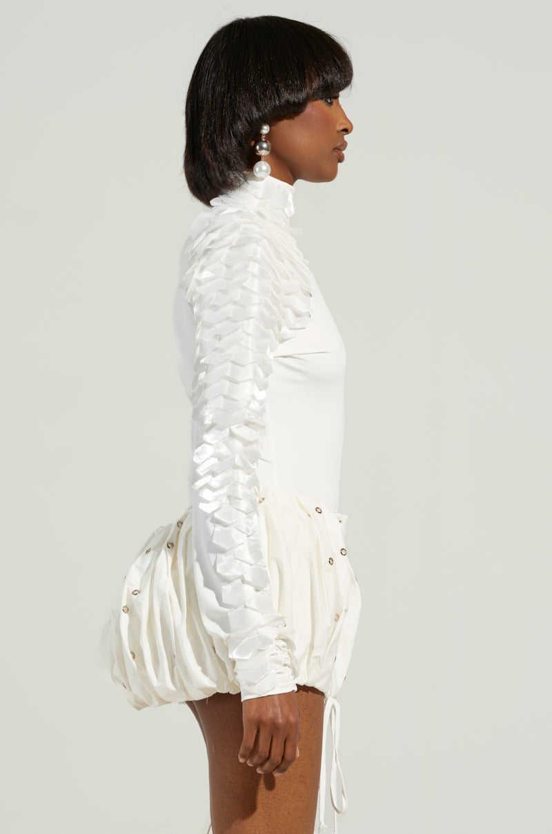 A person wearing the NADIA ORGANZA LONG SLEEVE SCALE TOP, featuring a mock neck design and textured ruffles, paired with a poufy skirt embellished with small metallic accents. Large pearl earrings complete the elegant look against a plain background that highlights the outfit's sophisticated charm.