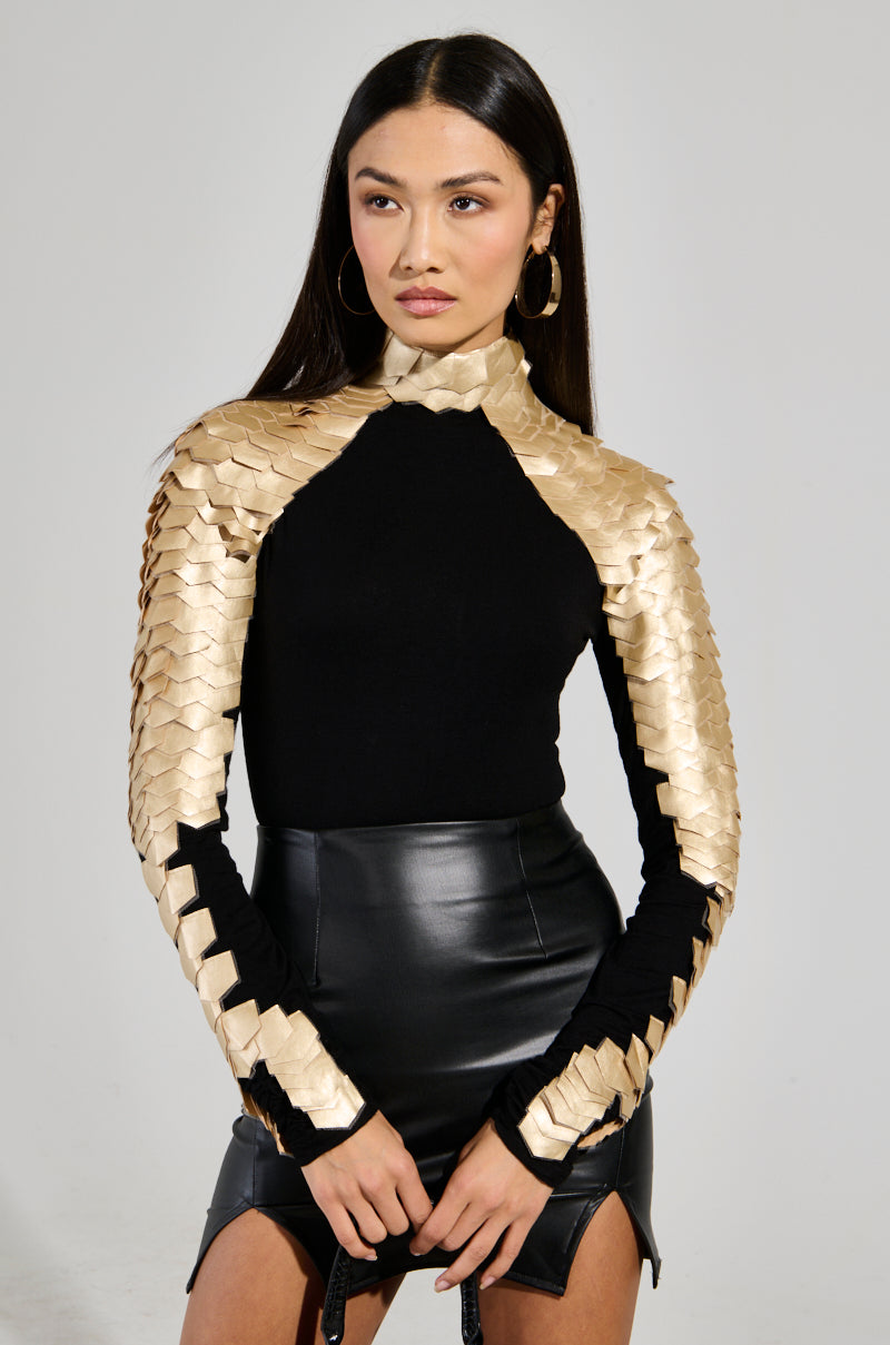 A woman stands confidently in the NASTASIA 2 Scale Top in gold, featuring scale-like detailing and a mock neck. Paired with a sleek faux leather skirt, her long straight hair and large hoop earrings enhance her bold look against a plain white background. 1