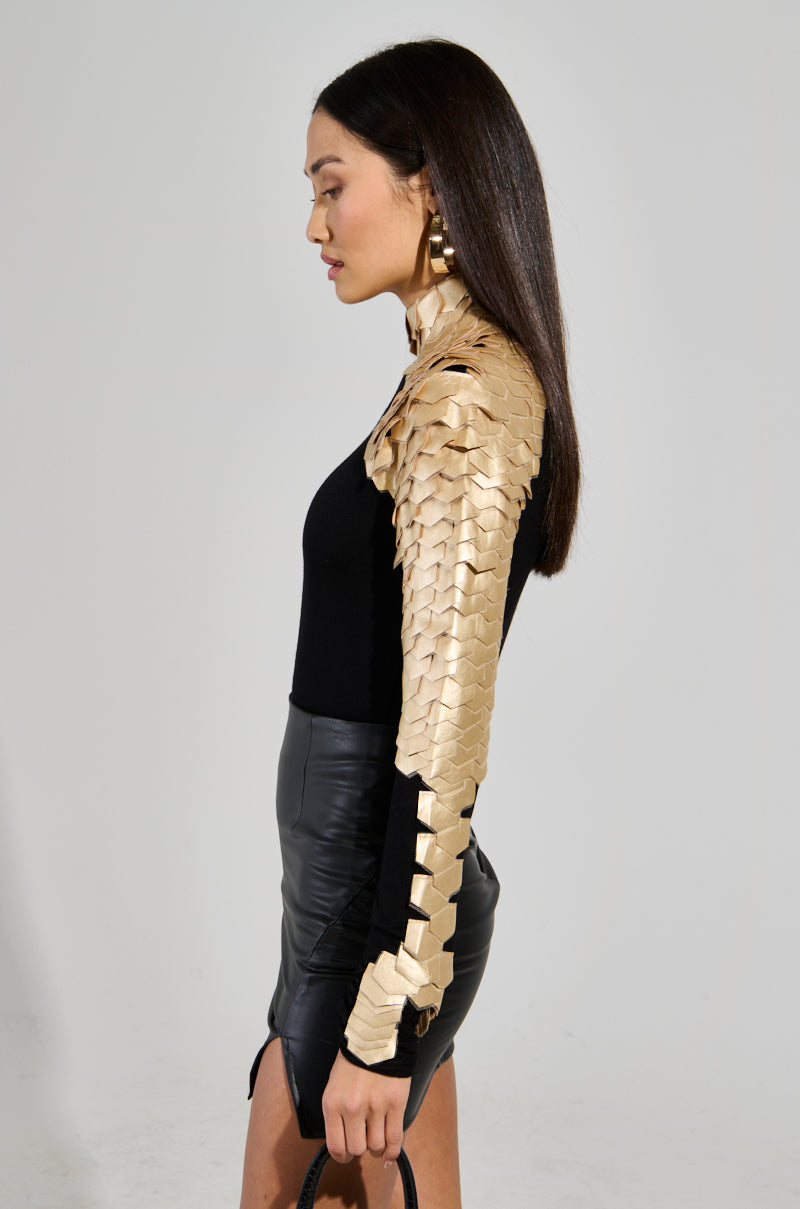 A person with long, straight hair is wearing the Nastasia 2 Scale Top in Gold, featuring a black and gold mock neck design with scale-like patterns on the shoulder and sleeve. They hold a black faux leather handbag, standing in profile against a plain white background. 3