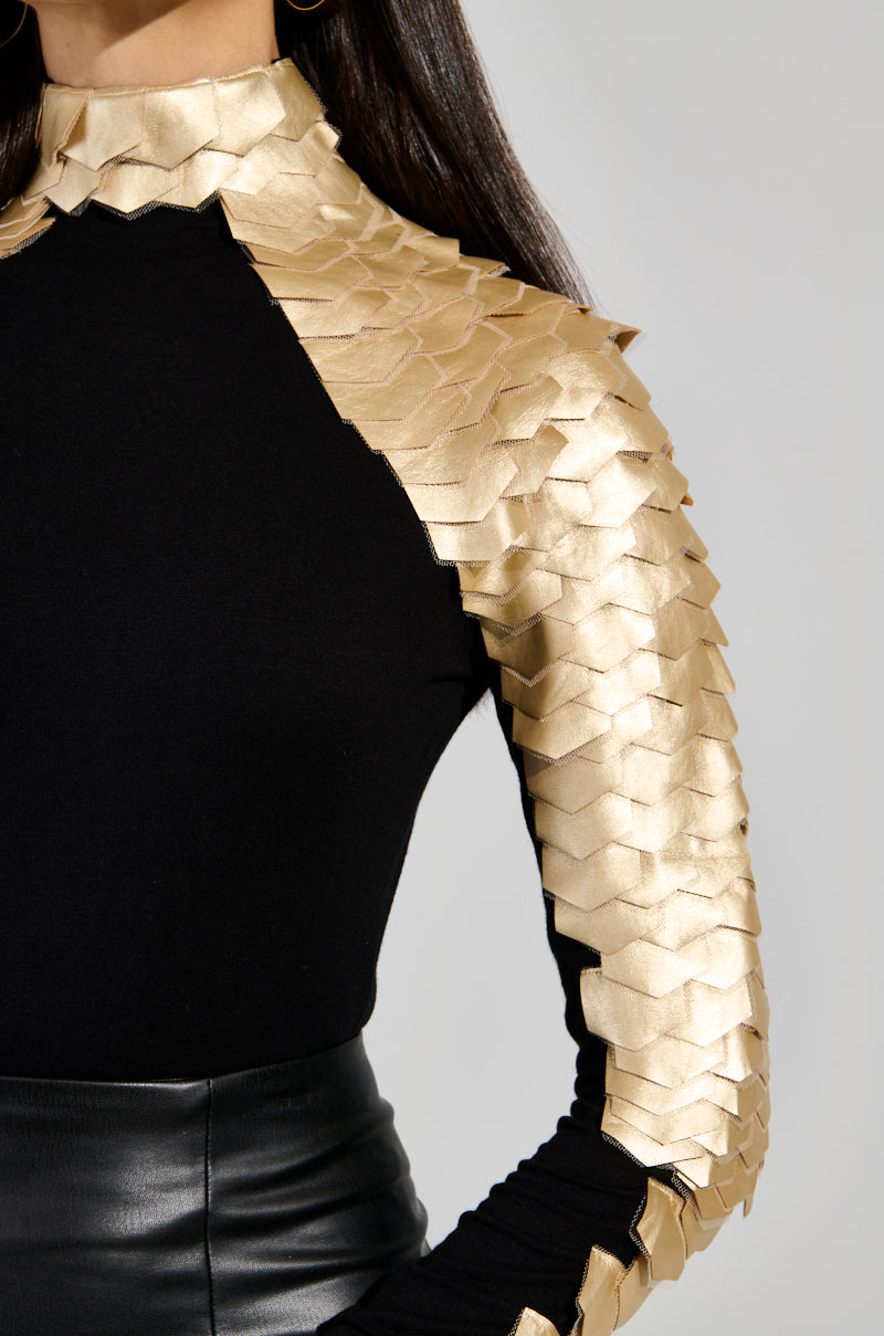 Someone wears the NASTASIA 2 SCALE TOP IN GOLD, featuring scale-like shoulder and sleeve embellishments, paired with a black faux leather skirt. The ensemble's mock neck adds sophistication against the neutral background. 6