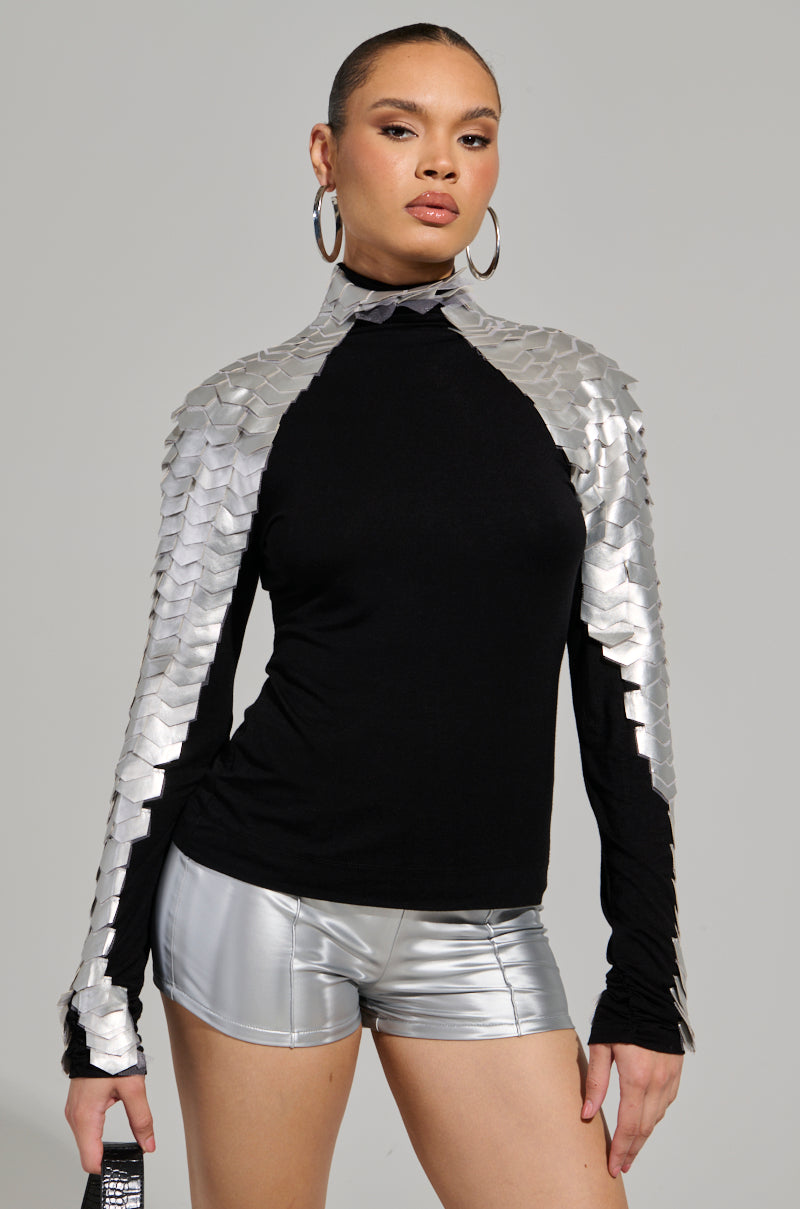 A person stands confidently, wearing the NASTASIA 2 SCALE TOP in black, featuring stretchy knit fabric with metallic silver scales on the shoulders and sleeves. They also flaunt shiny silver shorts, large hoop earrings, and hold a small black clutch. Their hair is pulled back neatly.