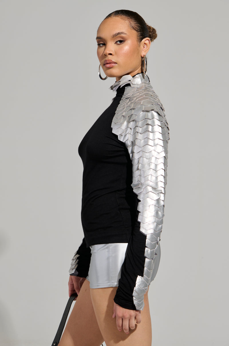 Confidently standing, a person wears the NASTASIA 2 SCALE TOP—a stretchy knit adorned with metallic silver scales on one sleeve. They accessorize with hoop earrings and hold a black stick, complementing the look with shiny silver shorts featuring ruched detailing against a neutral background.