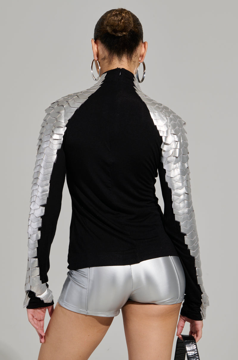 A person with short hair is turned away, wearing the NASTASIA 2 SCALE TOP, which features stretchy knit fabric and metallic silver scales on the shoulders and long sleeves. They are accessorized with large hoop earrings and shiny silver shorts while carrying a small bag.