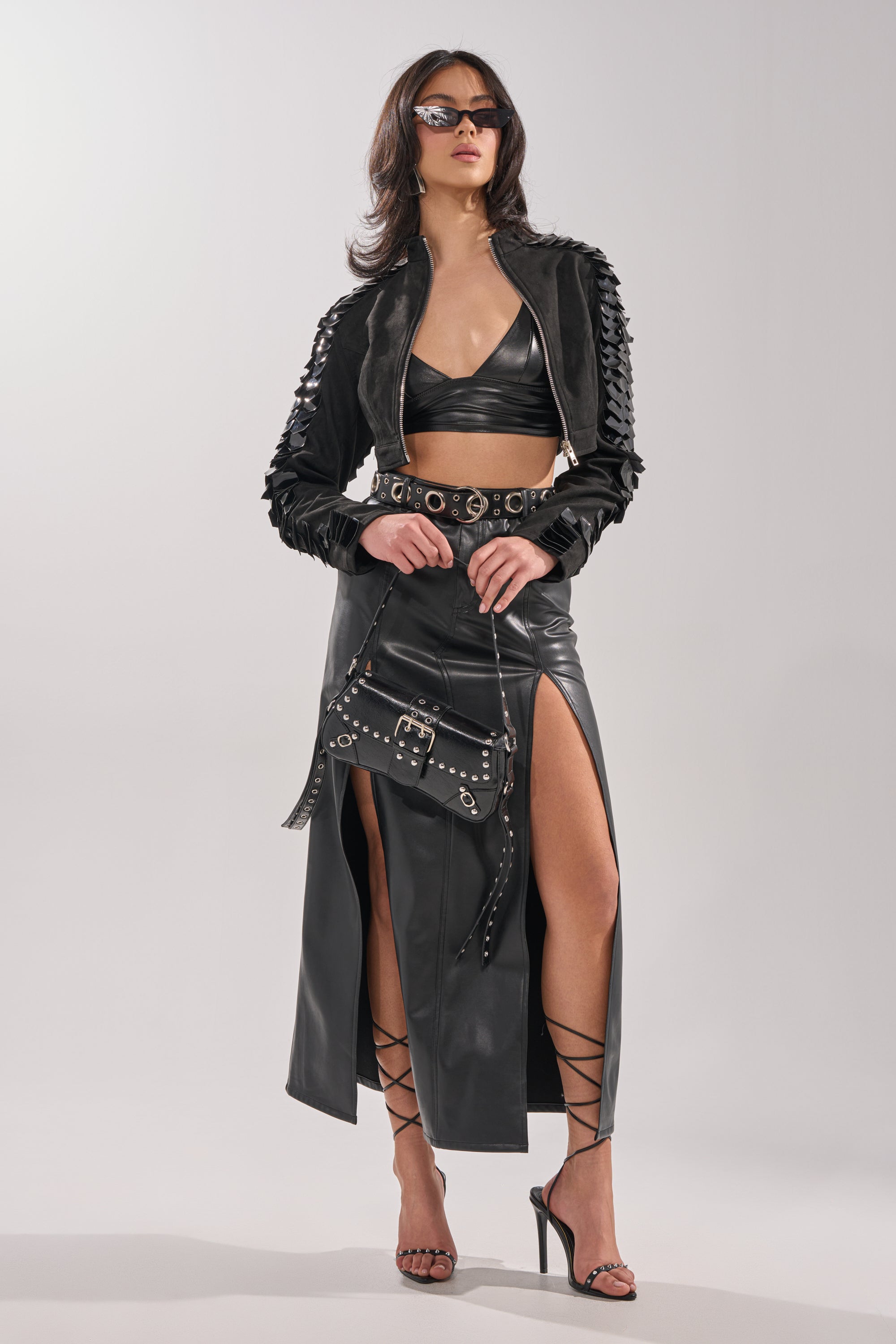 A woman stands against a plain background wearing the MOVING ON FAUX SUEDE MOTO jacket, sunglasses, a black cropped top with a mock neck, a high-slit black leather skirt, strappy heels, and holding a studded black handbag. 6