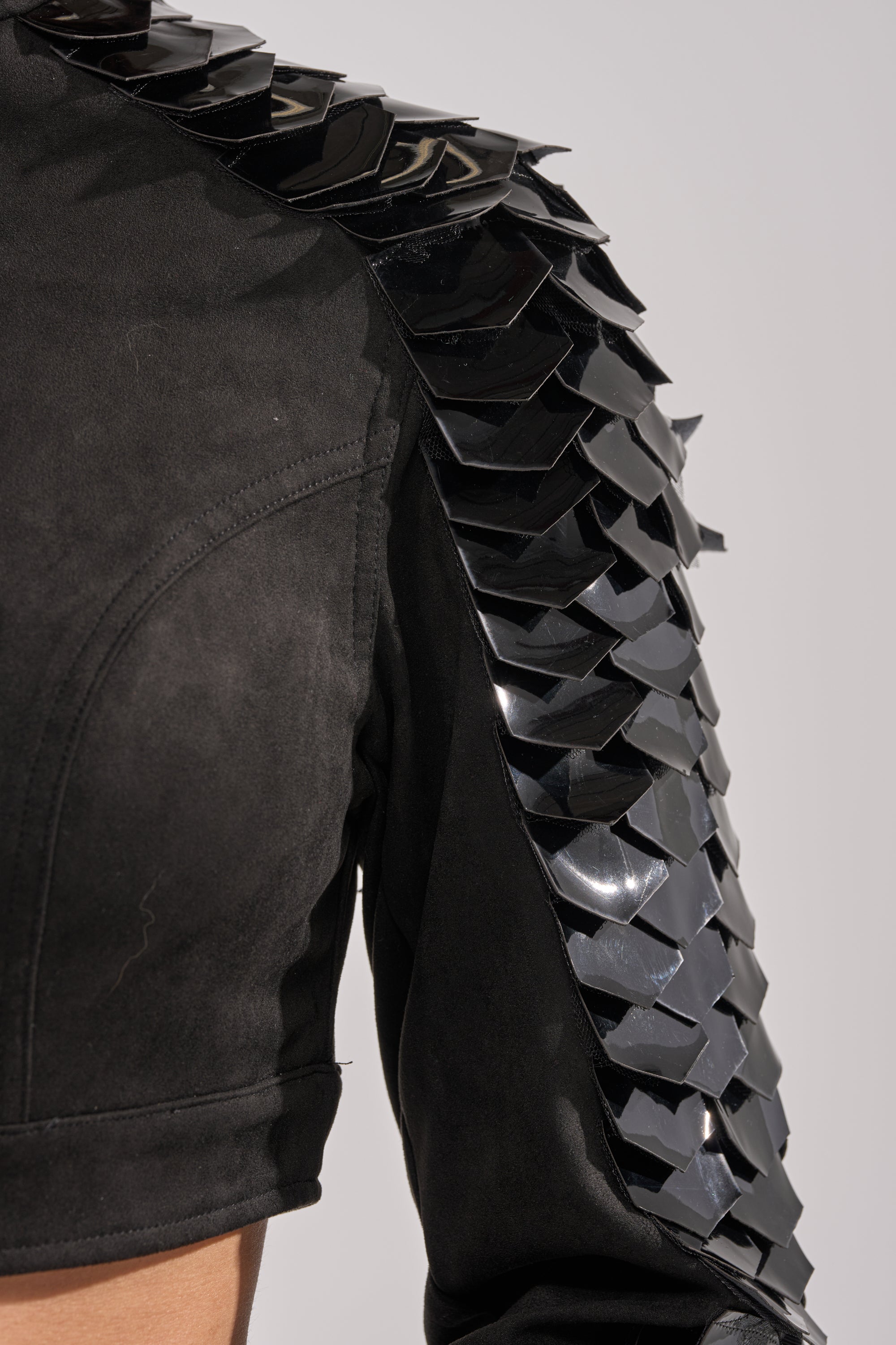 A close-up of the MOVING ON FAUX SUEDE MOTO jacket highlights shiny black scale-like embellishments on the shoulder and upper sleeve, giving it a textured, armored look against a plain light background. 7