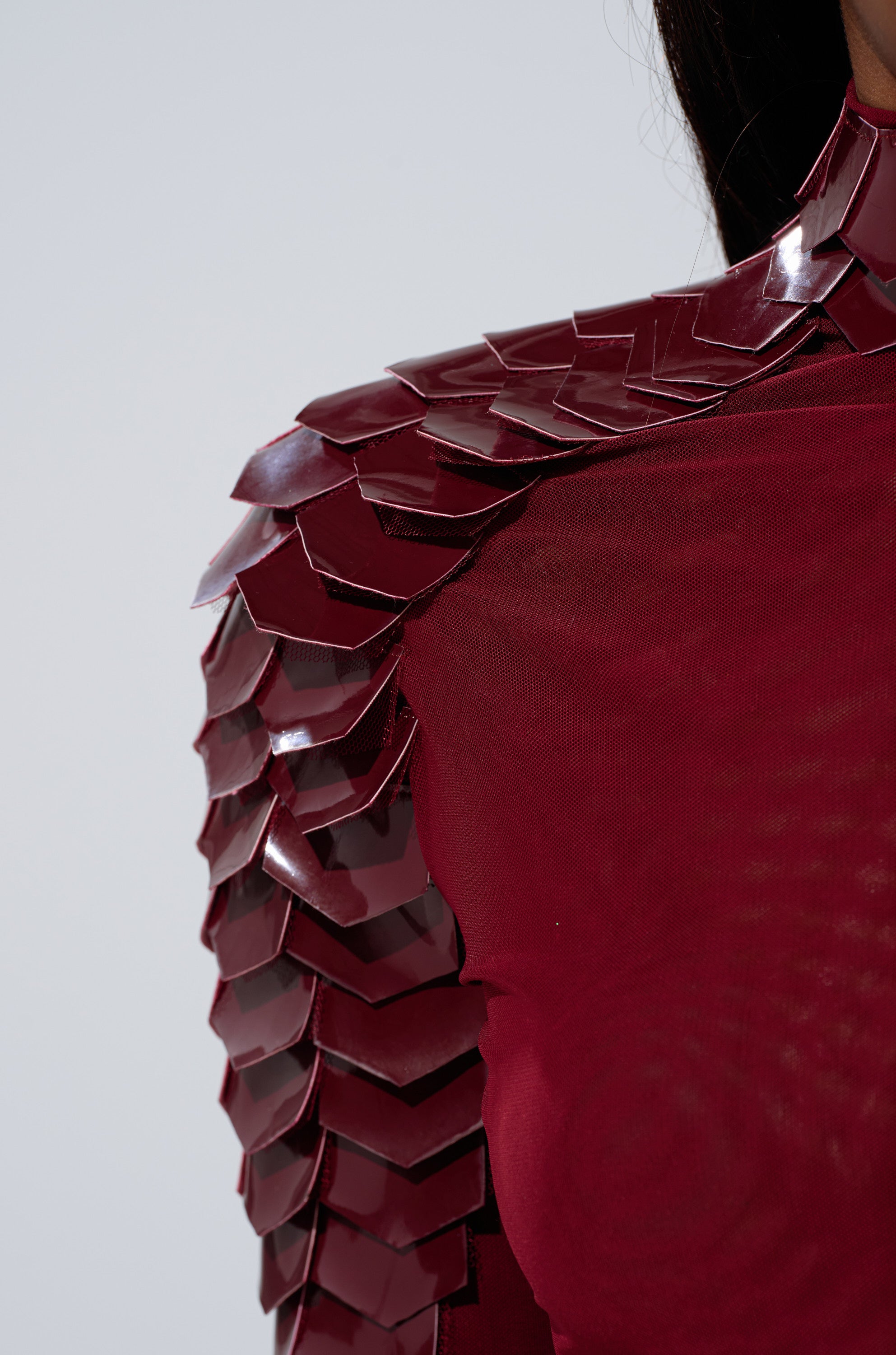 Close-up of a person wearing the VELMA VINYL SCALE TOP IN BURGUNDY, featuring shiny, layered scale-like embellishments on the shoulder and upper sleeve, set against a light grey background.