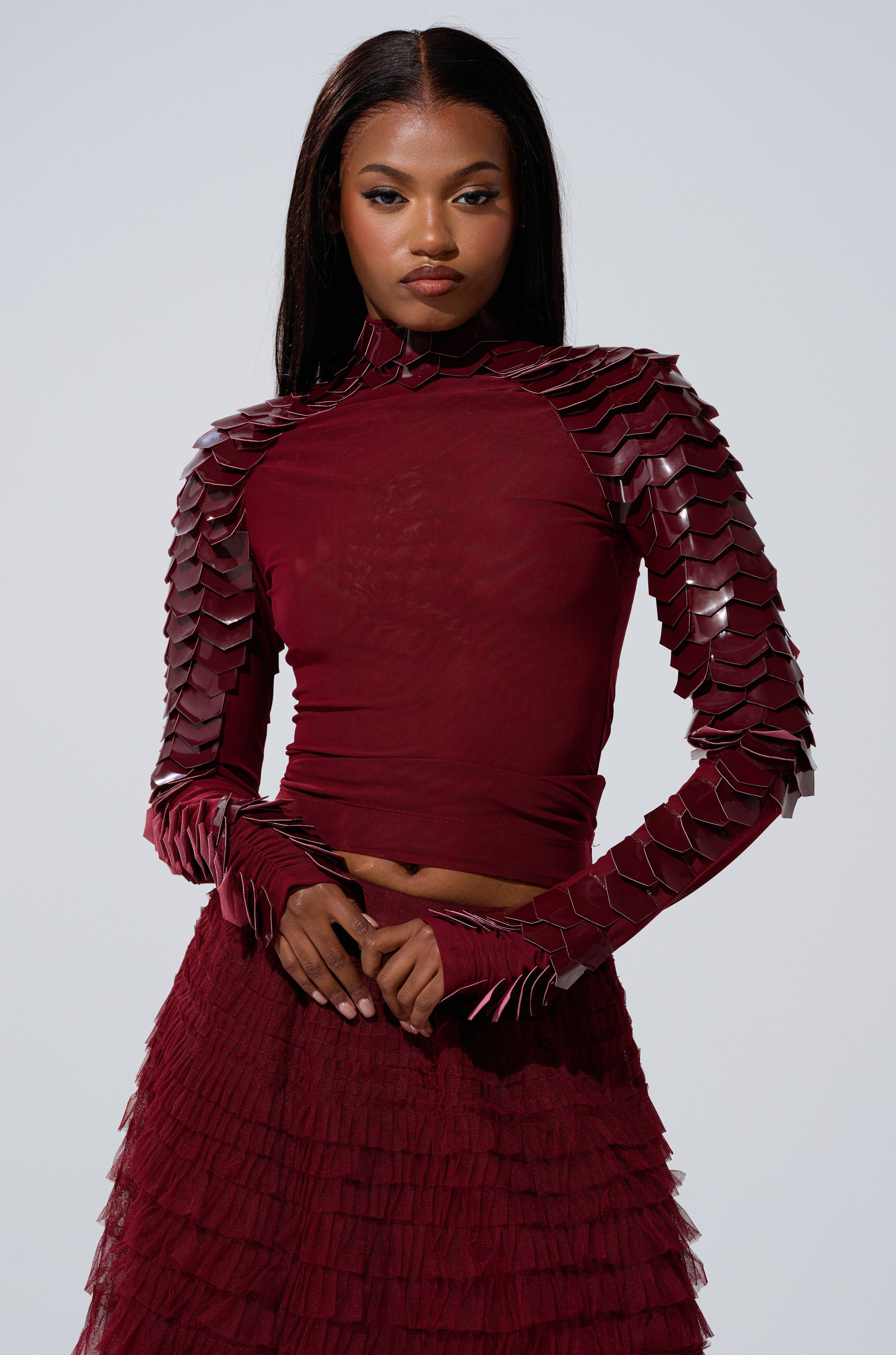 A woman in a fitted VELMA VINYL SCALE TOP IN BURGUNDY with sculpted, layered shoulder and arm details, paired with a matching textured skirt, stands confidently against a plain light background.