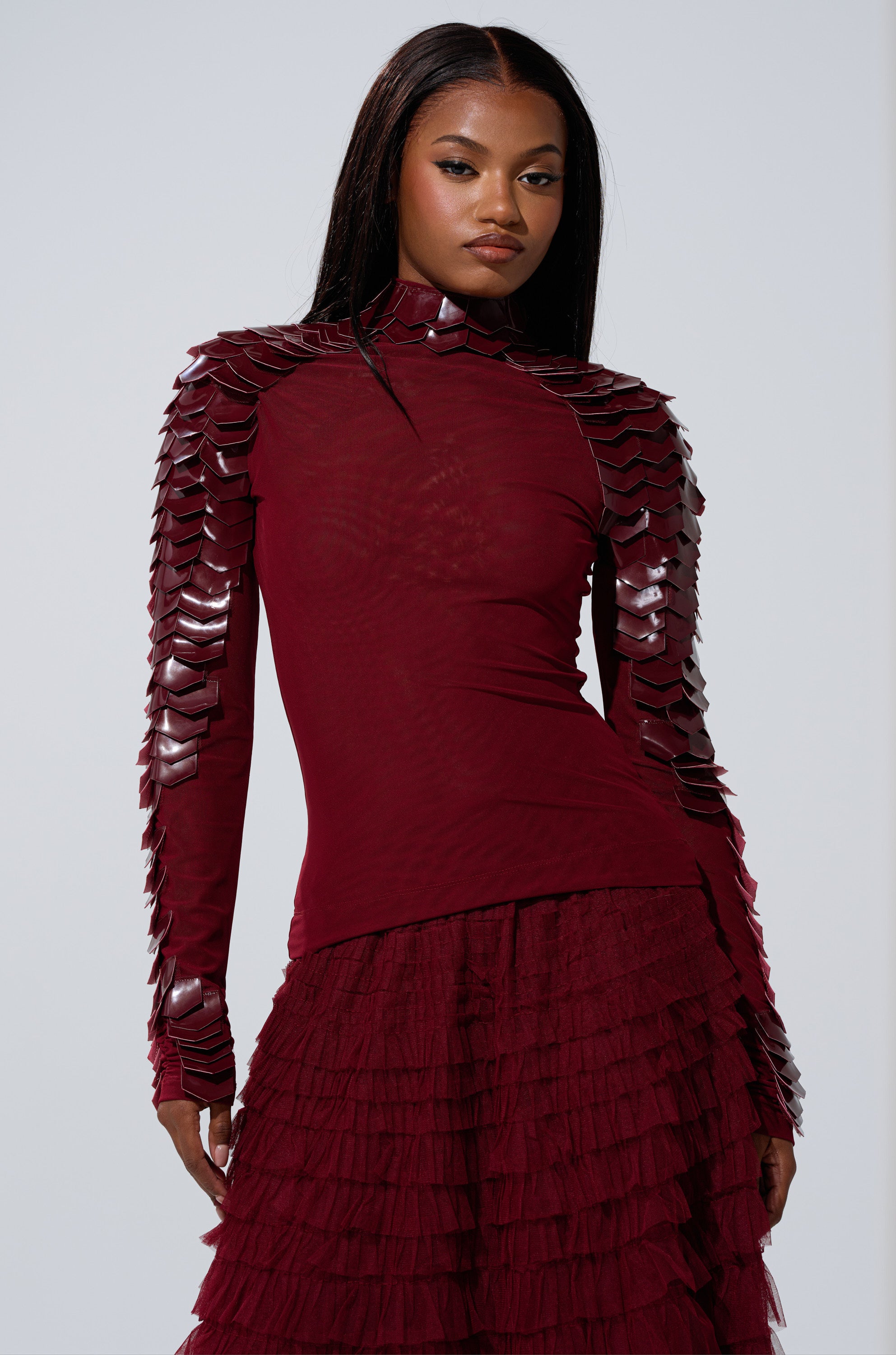 A woman wearing the VELMA VINYL SCALE TOP IN BURGUNDY—a fitted mesh top with shiny scale-like embellishments—paired with a matching burgundy ruffled skirt, stands against a plain light grey background.