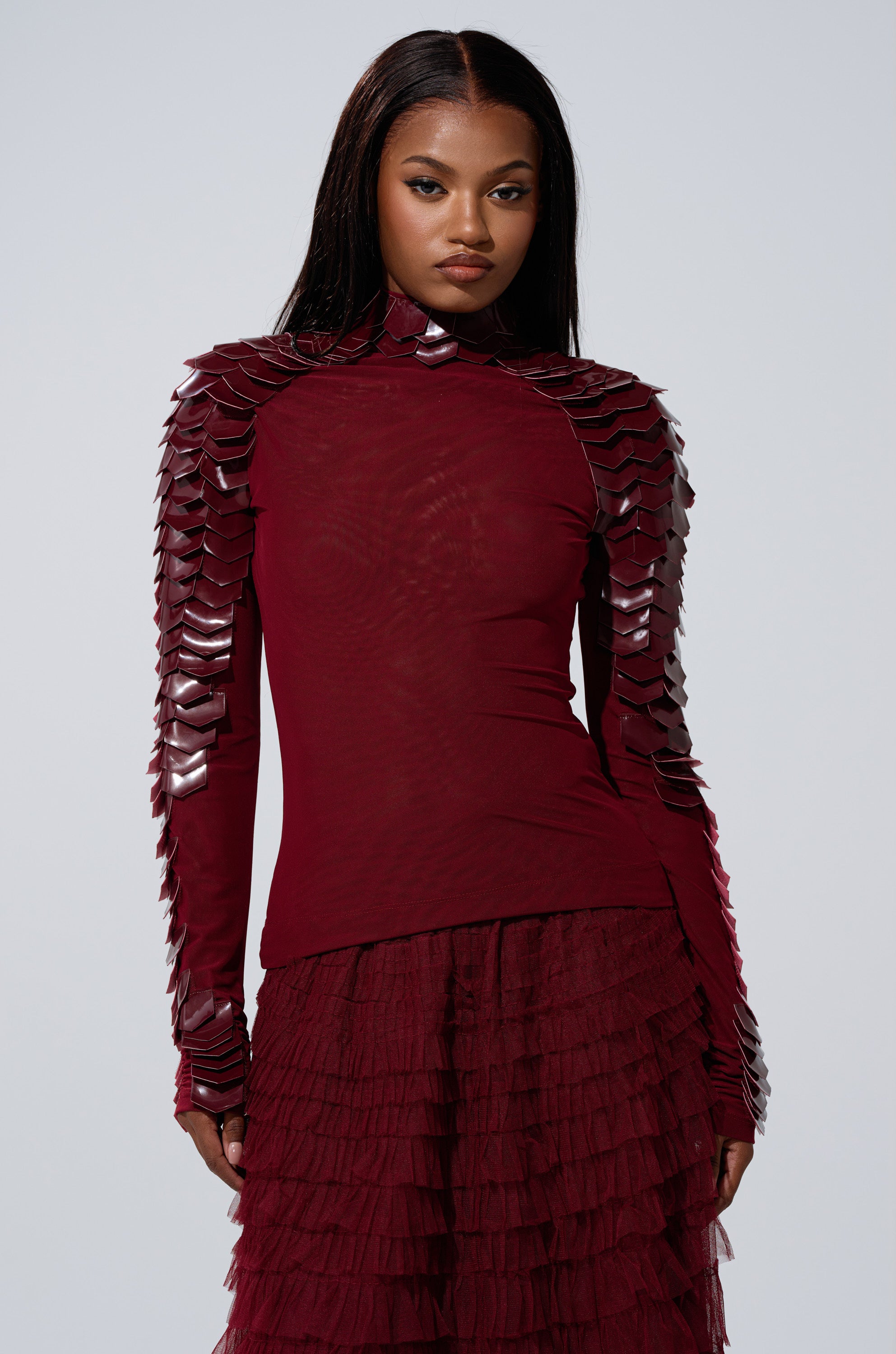 A woman wears the VELMA VINYL SCALE TOP IN BURGUNDY, featuring long sleeves and scale-like details, paired with a matching ruffled skirt, as she stands against a plain light background.