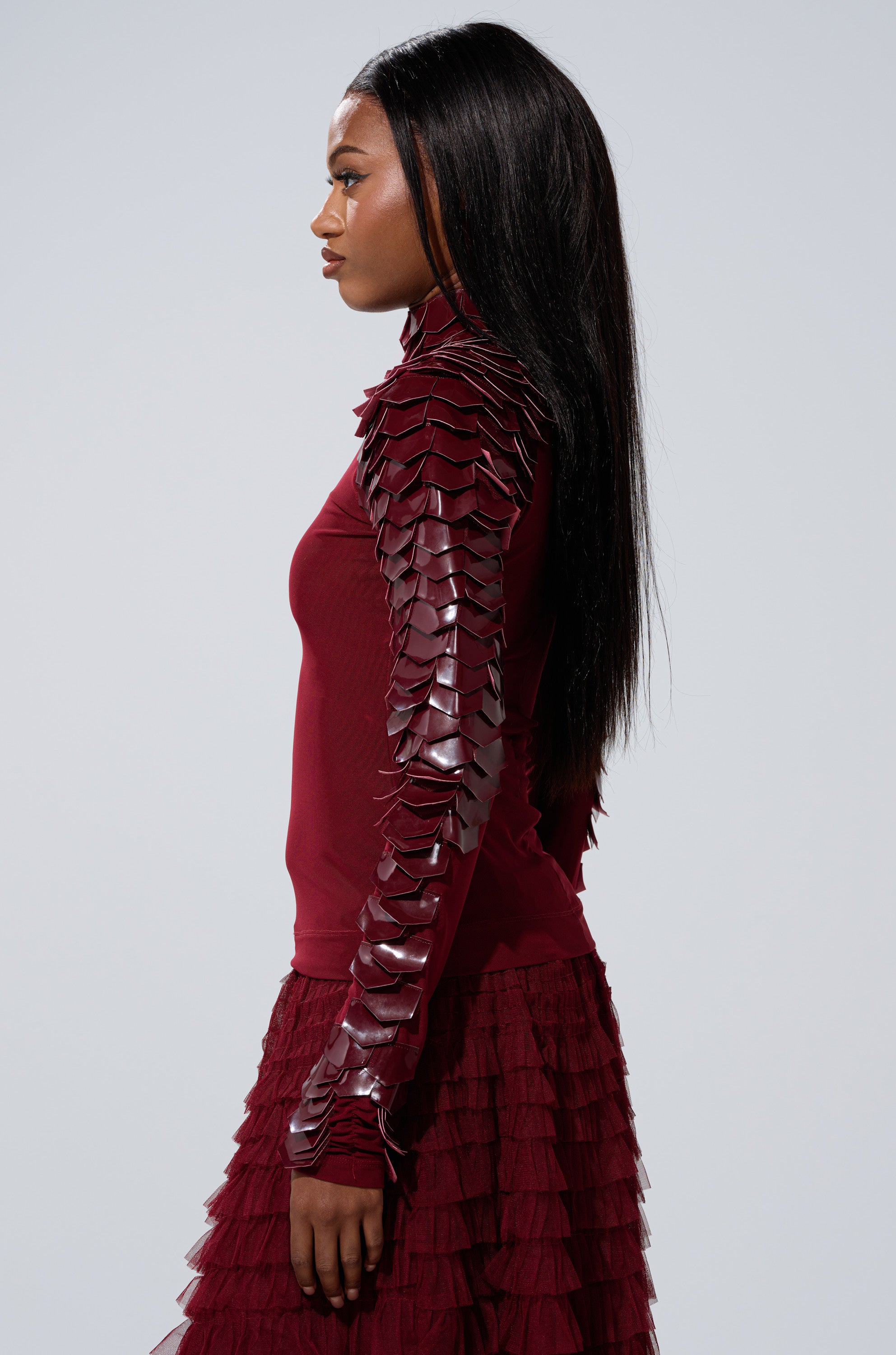 A woman stands in profile wearing the VELMA VINYL SCALE TOP IN BURGUNDY, a curve-hugging mesh top with layered, scale-like sleeve details. She has long, straight hair and a neutral expression against a plain background.