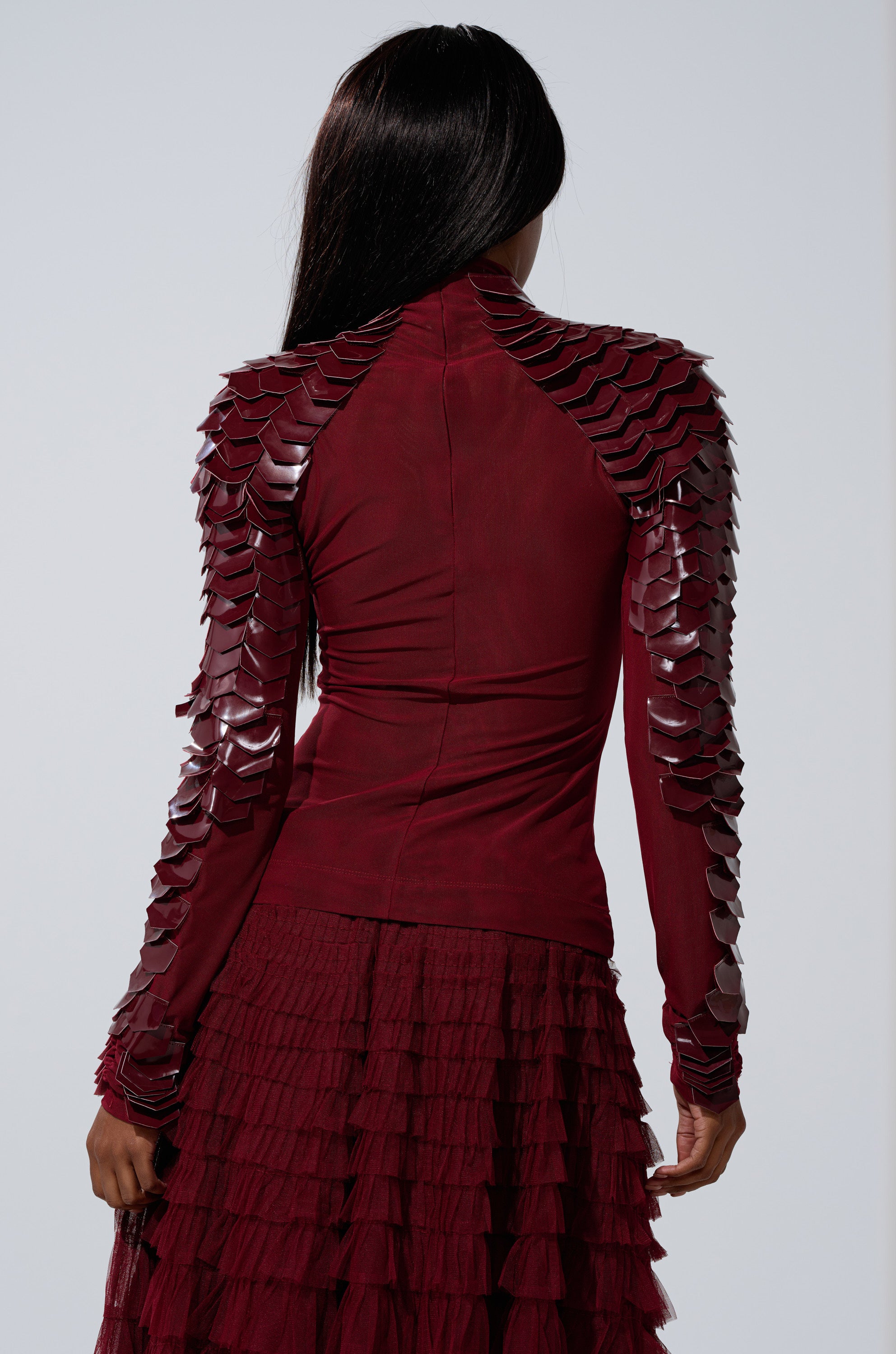 A person with long dark hair, seen from the back, wears the VELMA VINYL SCALE TOP IN BURGUNDY, featuring shiny scale-like shoulder details and long sleeves, styled with a curve-hugging ruffled maroon skirt.