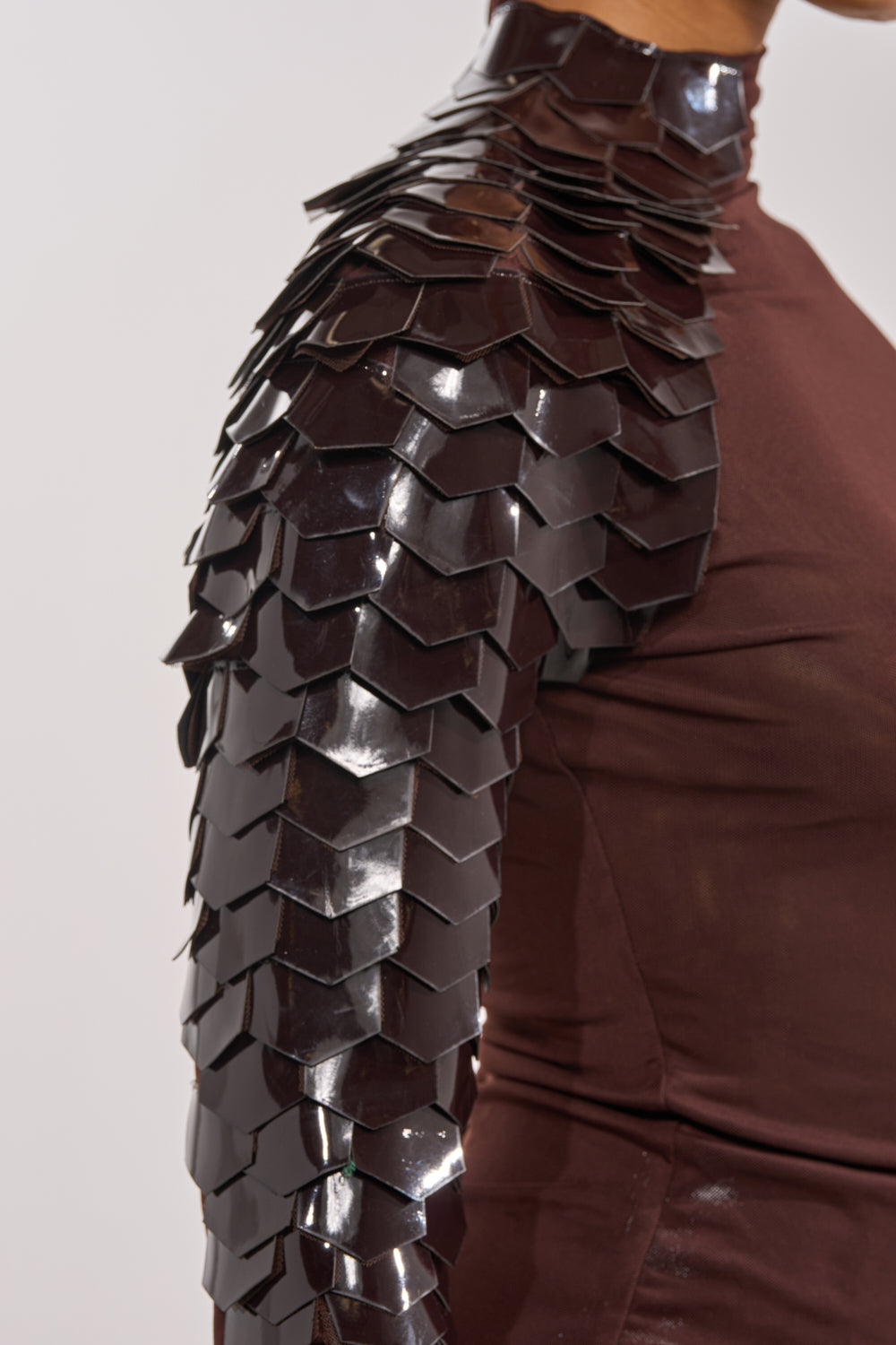 A close-up of a person's upper body shows the VELMA VINYL SINGLE SLEEVE SCALE TOP IN BROWN, featuring a fitted brown mesh with glossy black armor-like scales and a faux leather sleeve for an avant-garde look. 9