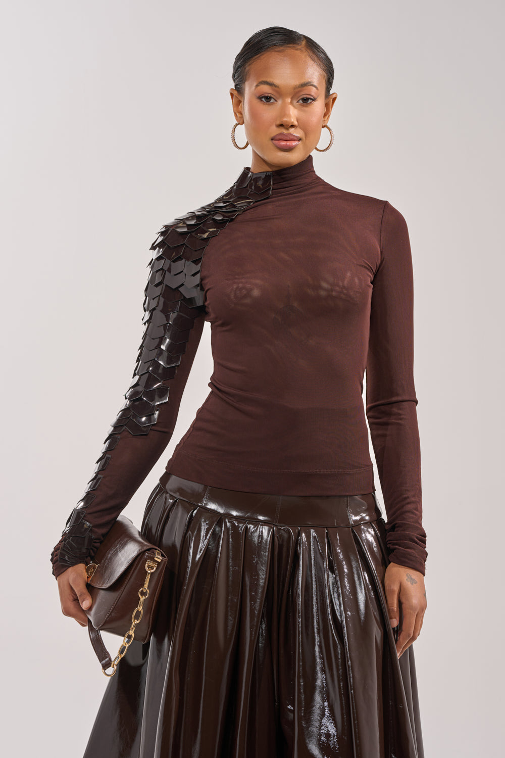 A woman wears the VELMA VINYL SINGLE SLEEVE SCALE TOP IN BROWN, styled with a glossy brown high-waisted skirt, gold chain brown handbag, and hoop earrings for an avant-garde look. 2