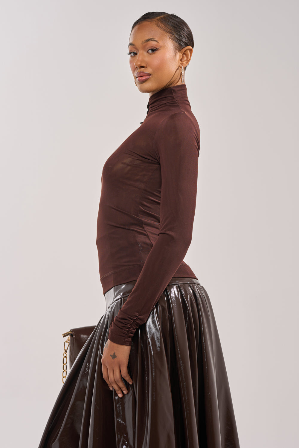 A woman in the VELMA VINYL SINGLE SLEEVE SCALE TOP IN BROWN and a shiny black skirt stands in profile, holding a clutch with a gold chain against a plain light background. 4