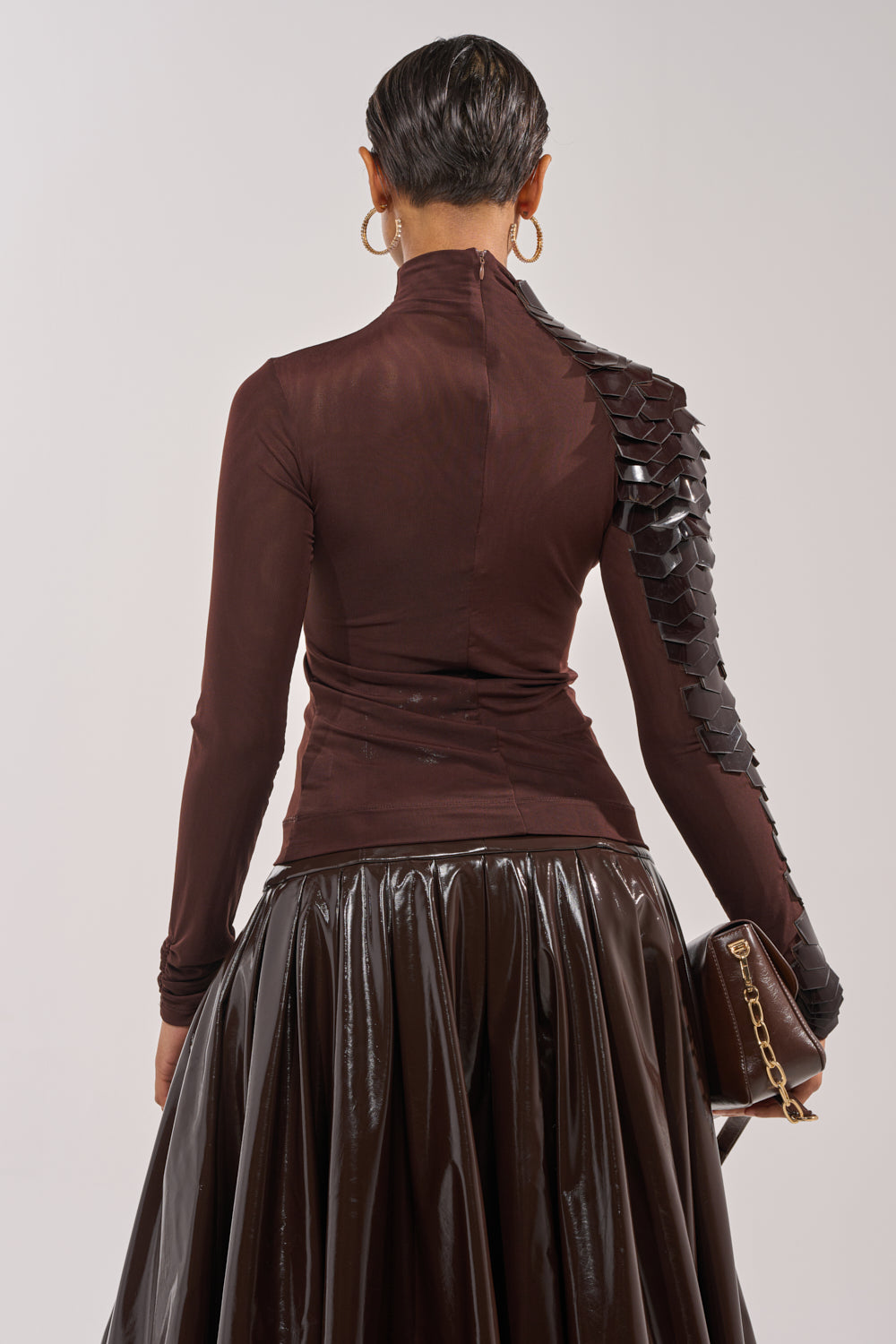 A person with short hair and large hoop earrings wears the VELMA VINYL SINGLE SLEEVE SCALE TOP IN BROWN, featuring a fitted design with faux leather and black textured detail, paired with a shiny brown skirt and a brown clutch with gold chain. 5
