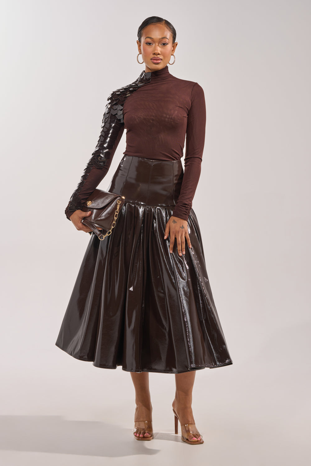 A woman wears the VELMA VINYL SINGLE SLEEVE SCALE TOP IN BROWN with a dark brown midi skirt, clear heeled sandals, gold hoop earrings, and holds a matching clutch. 6