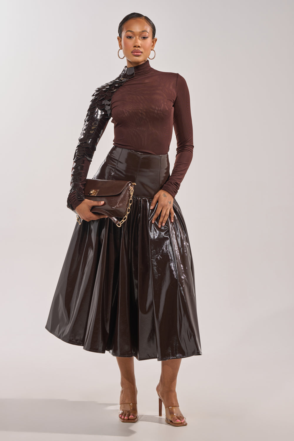 A woman stands against a plain background wearing a shiny brown midi skirt, the VELMA VINYL SINGLE SLEEVE SCALE TOP IN BROWN, clear high heels, and holding a brown clutch with a gold chain. 7