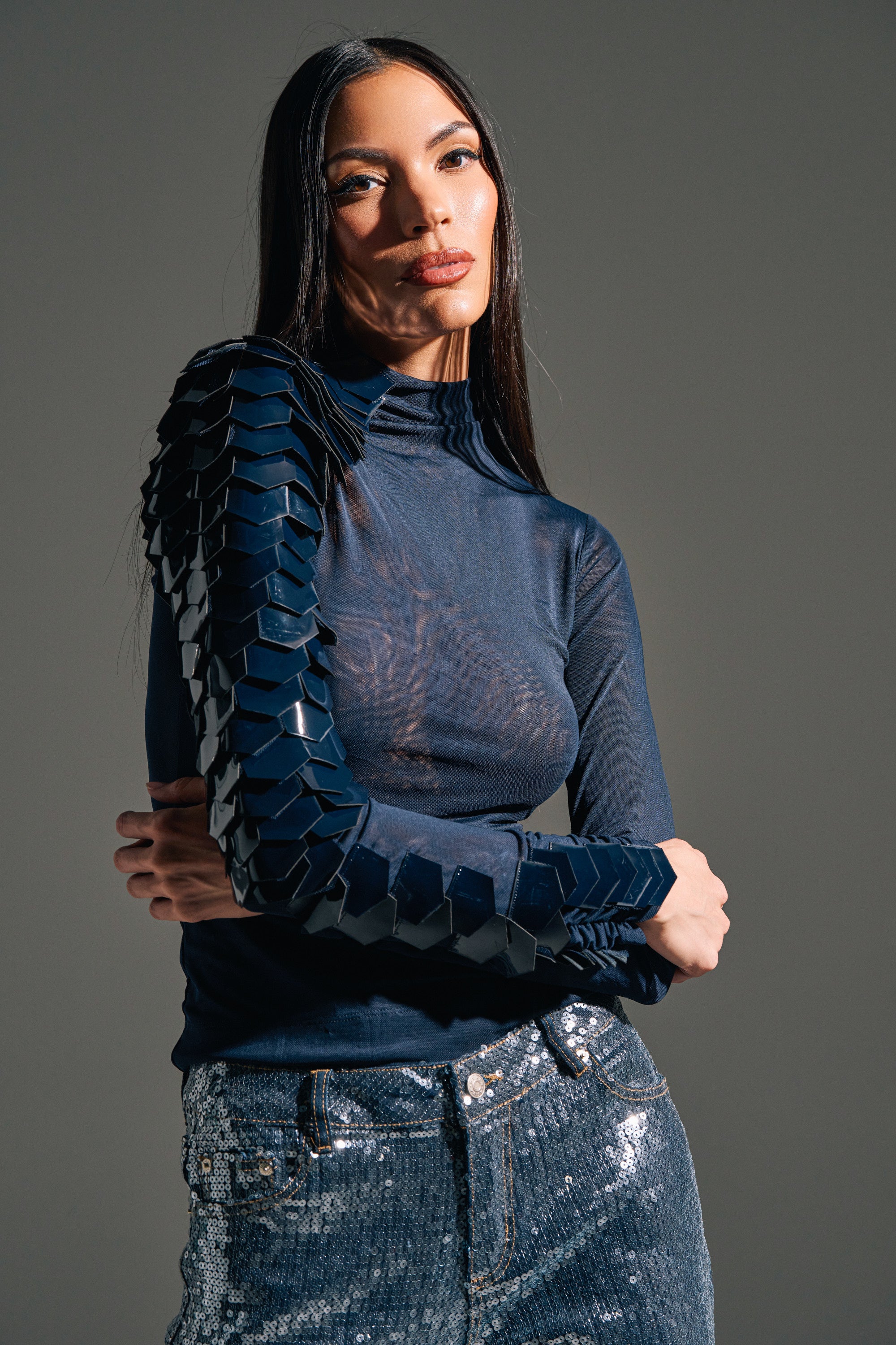 A woman with long dark hair wears the VELMA VINYL SINGLE SLEEVE SCALE TOP IN NAVY, featuring a sheer mesh high neck and 3D scale embellishments on one sleeve, paired with sequined jeans, standing against a gray background.