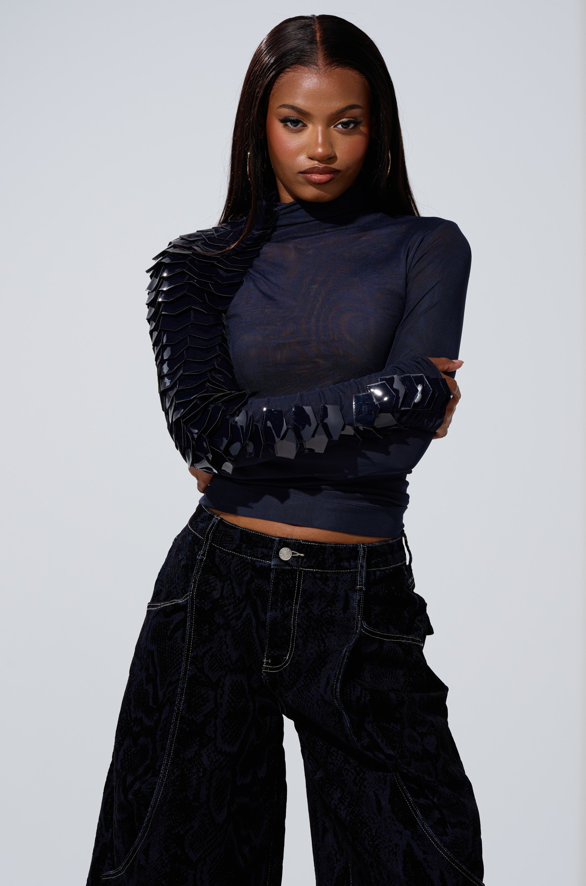 A woman with long straight hair wears the VELMA VINYL SINGLE SLEEVE SCALE TOP IN NAVY featuring shiny, sculptural details and loose black pants. She stands with arms crossed against a plain light background, gazing confidently at the camera.