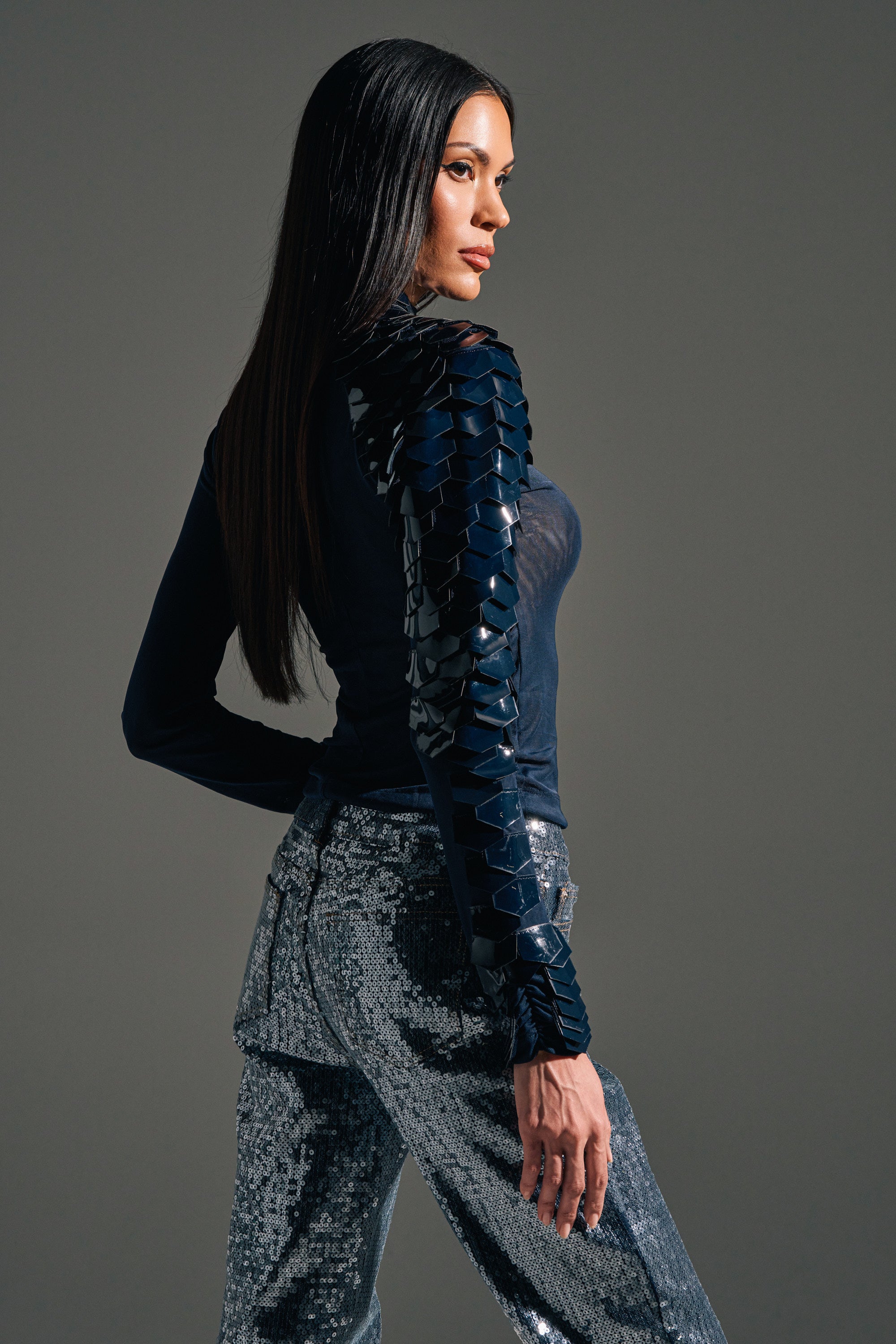 A woman with long straight hair stands in profile, wearing the Velma Vinyl Single Sleeve Scale Top in navy with glossy, armored detailing on one sleeve and silver sequin pants, set against a plain gray background.