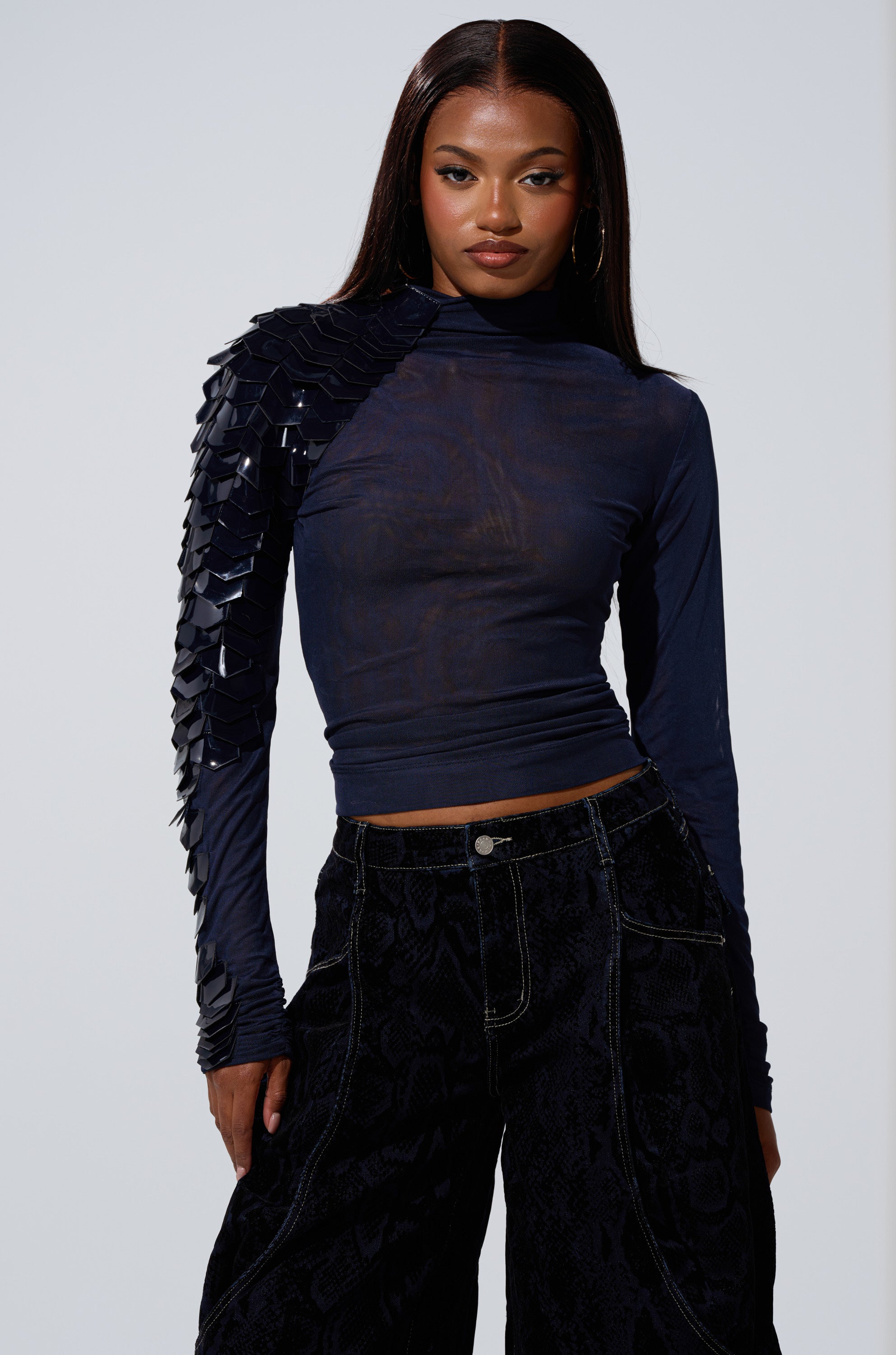 A woman wearing the VELMA VINYL SINGLE SLEEVE SCALE TOP IN NAVY with shiny black embellishments on one shoulder and black high-waisted pants, standing against a light background.