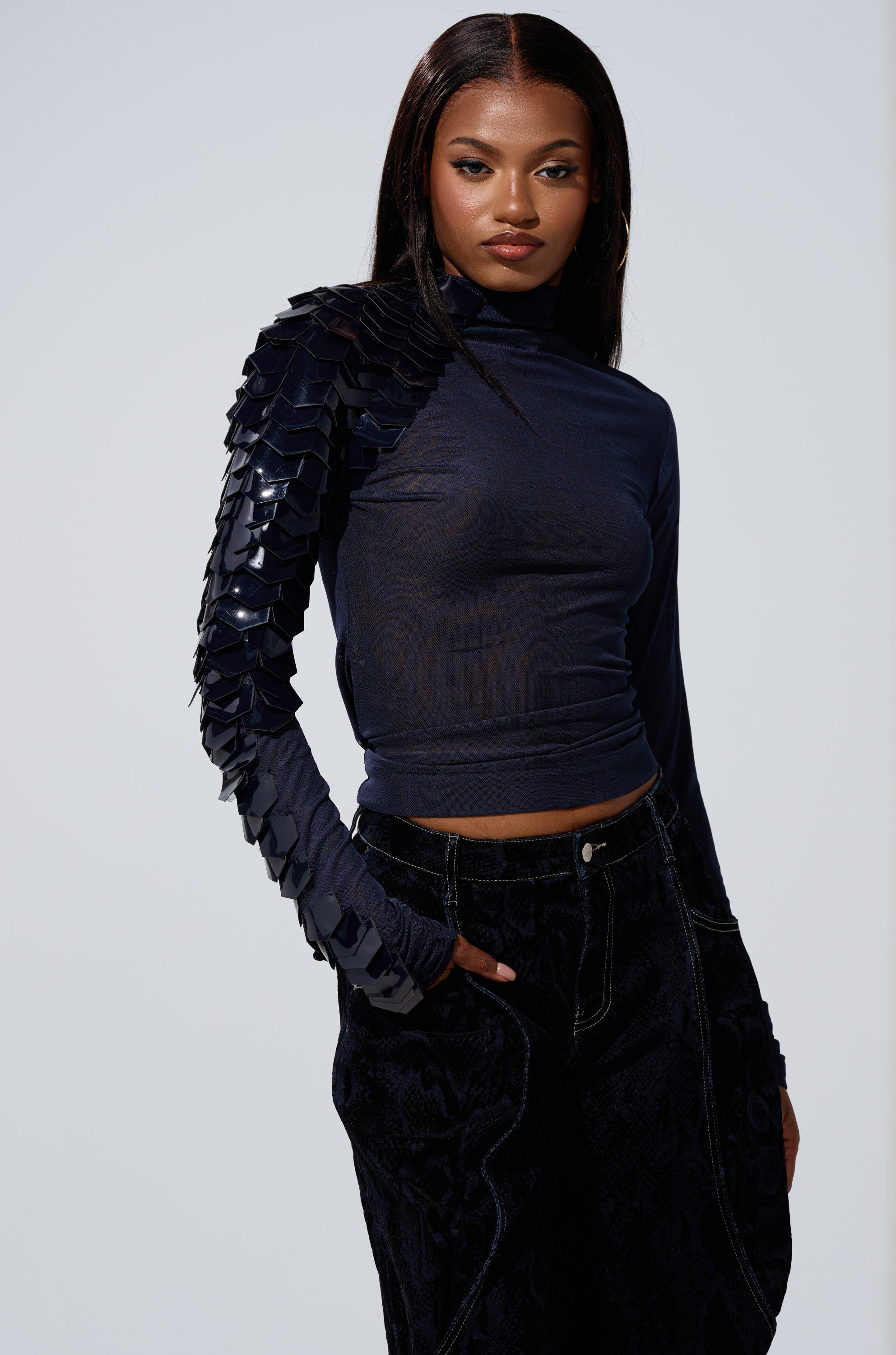 A woman with long straight hair models the VELMA VINYL SINGLE SLEEVE SCALE TOP IN NAVY, featuring shiny layered details on one sleeve, paired with black textured pants, posing confidently against a plain light background.