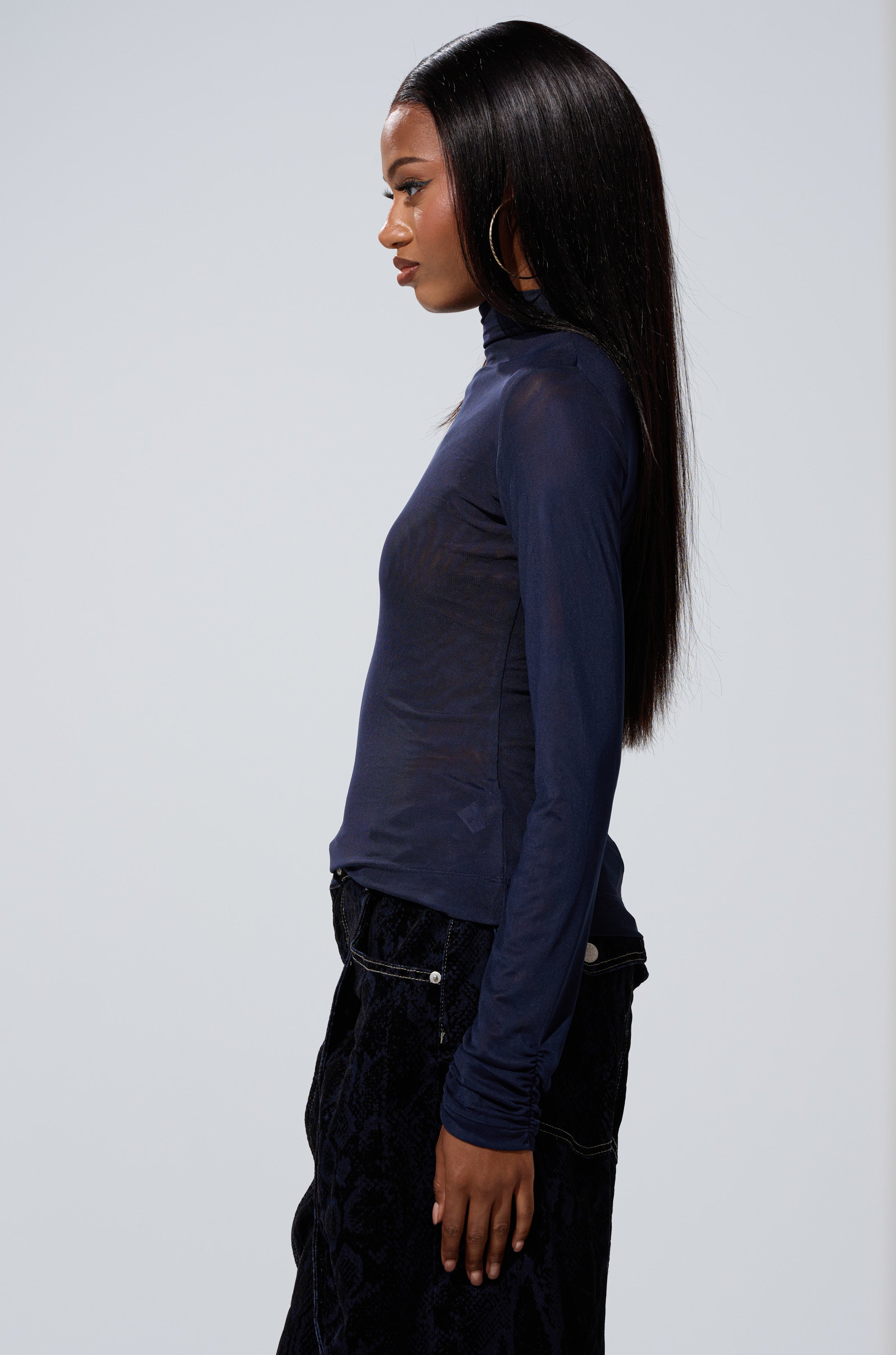 A woman with long straight hair stands in profile, wearing the VELMA VINYL SINGLE SLEEVE SCALE TOP IN NAVY and dark pants with zipper details, set against a plain light background.