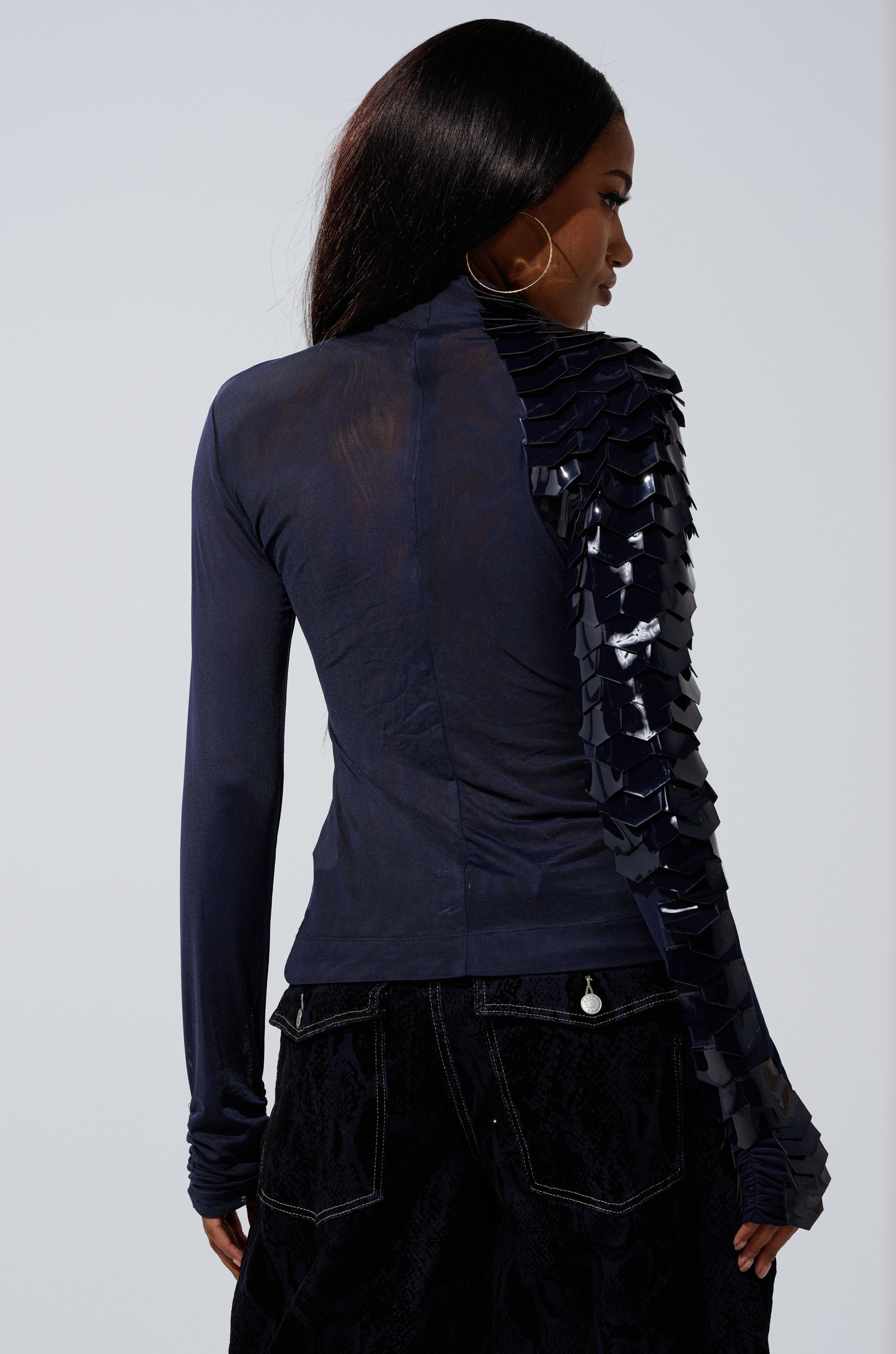 A woman stands facing away, wearing the VELMA VINYL SINGLE SLEEVE SCALE TOP IN NAVY, featuring shiny textured embellishments on one sleeve, paired with dark textured pants. Her long straight hair and large hoop earrings complete the look.
