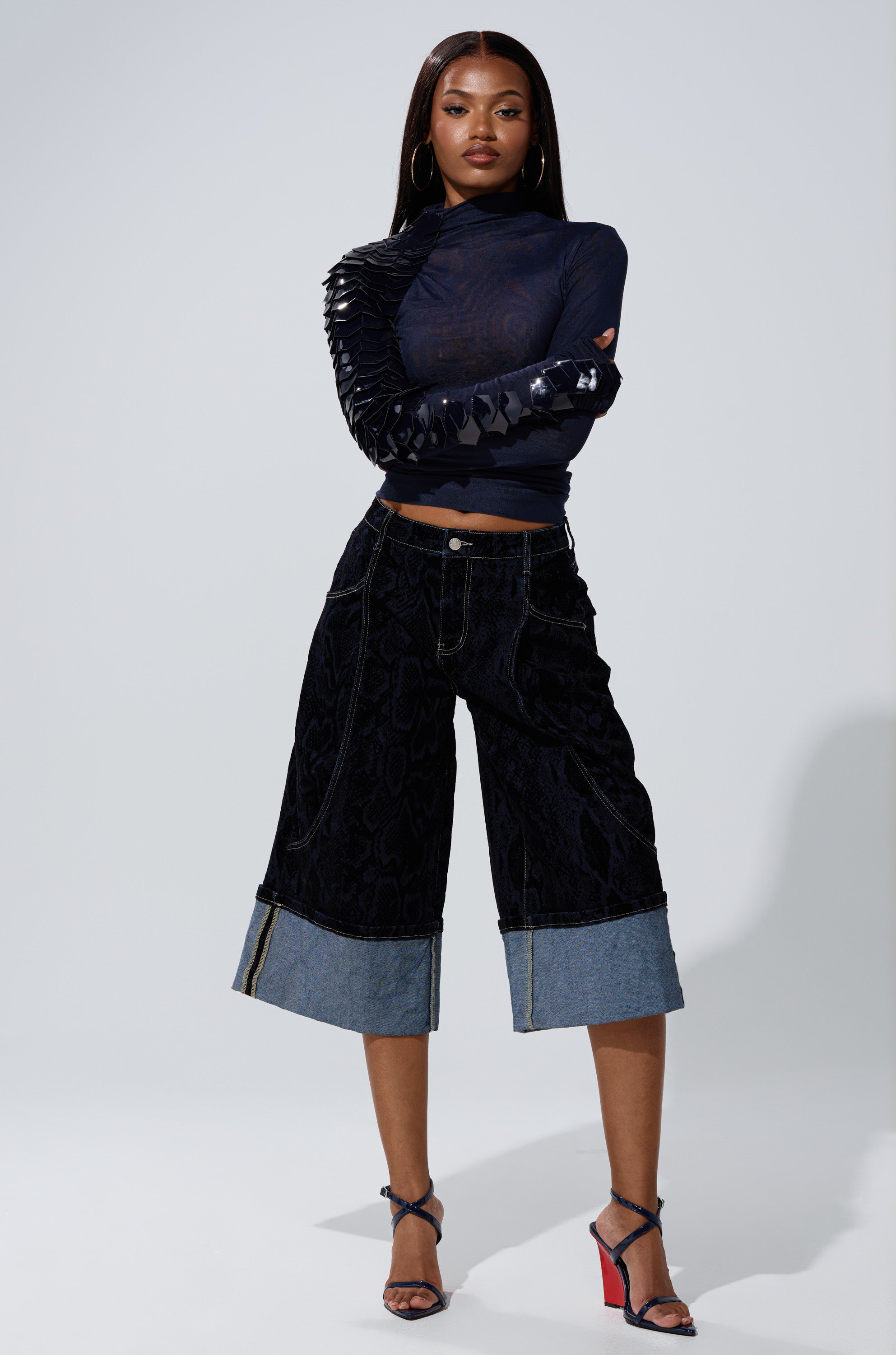 A woman stands confidently in the AKIRA Label VELMA VINYL SINGLE SLEEVE SCALE TOP IN NAVY, paired with knee-length wide-leg black denim pants and strappy high-heeled sandals, posing against a plain background.