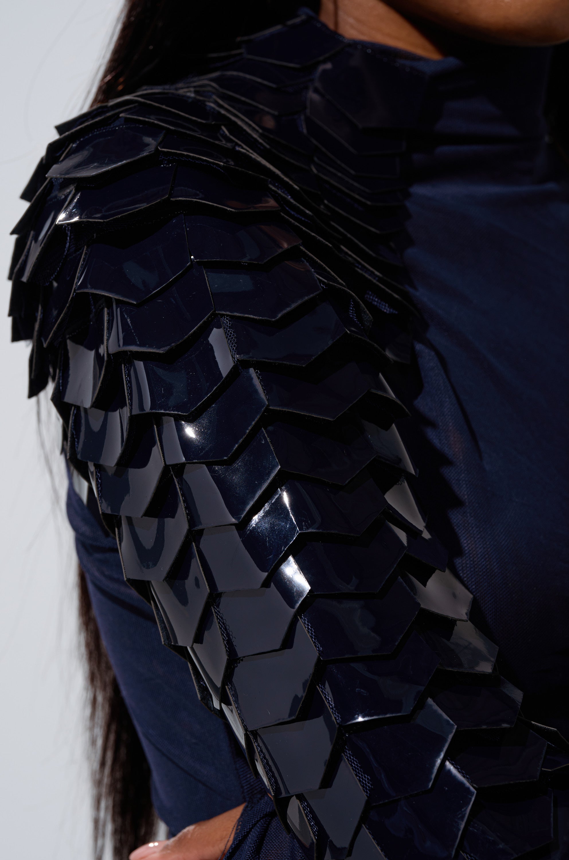 A close-up of a person wearing the VELMA VINYL SINGLE SLEEVE SCALE TOP IN NAVY, featuring dark, shiny, scale-like textured embellishments on one shoulder and sleeve, giving an armor-inspired look against a plain background.