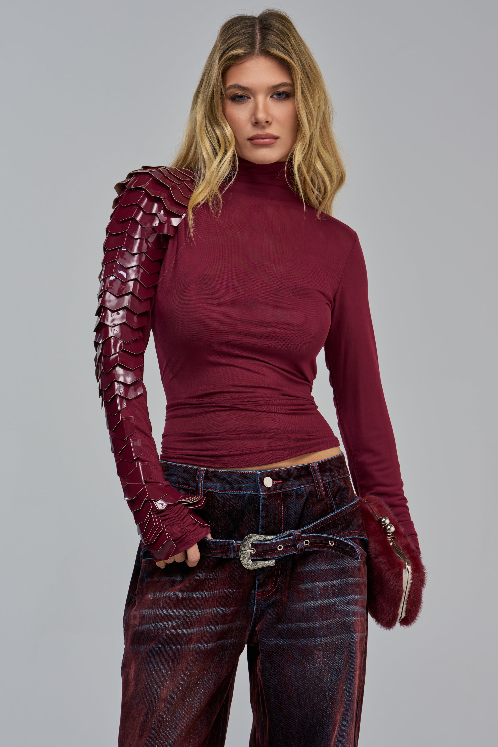 A person wears the VELMA VINYL SINGLE SLEEVE SCALE TOP IN PLUM, featuring a single vinyl scale-patterned sleeve on the right arm paired with dark, distressed jeans.