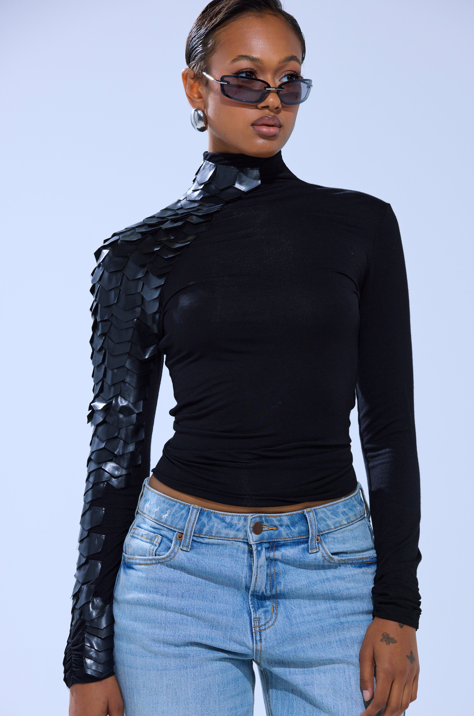 A person wearing sunglasses, the AKIRA Label NASTASIA SCALE TOP in black with shiny details on one sleeve, and light blue jeans stands against a plain light background.