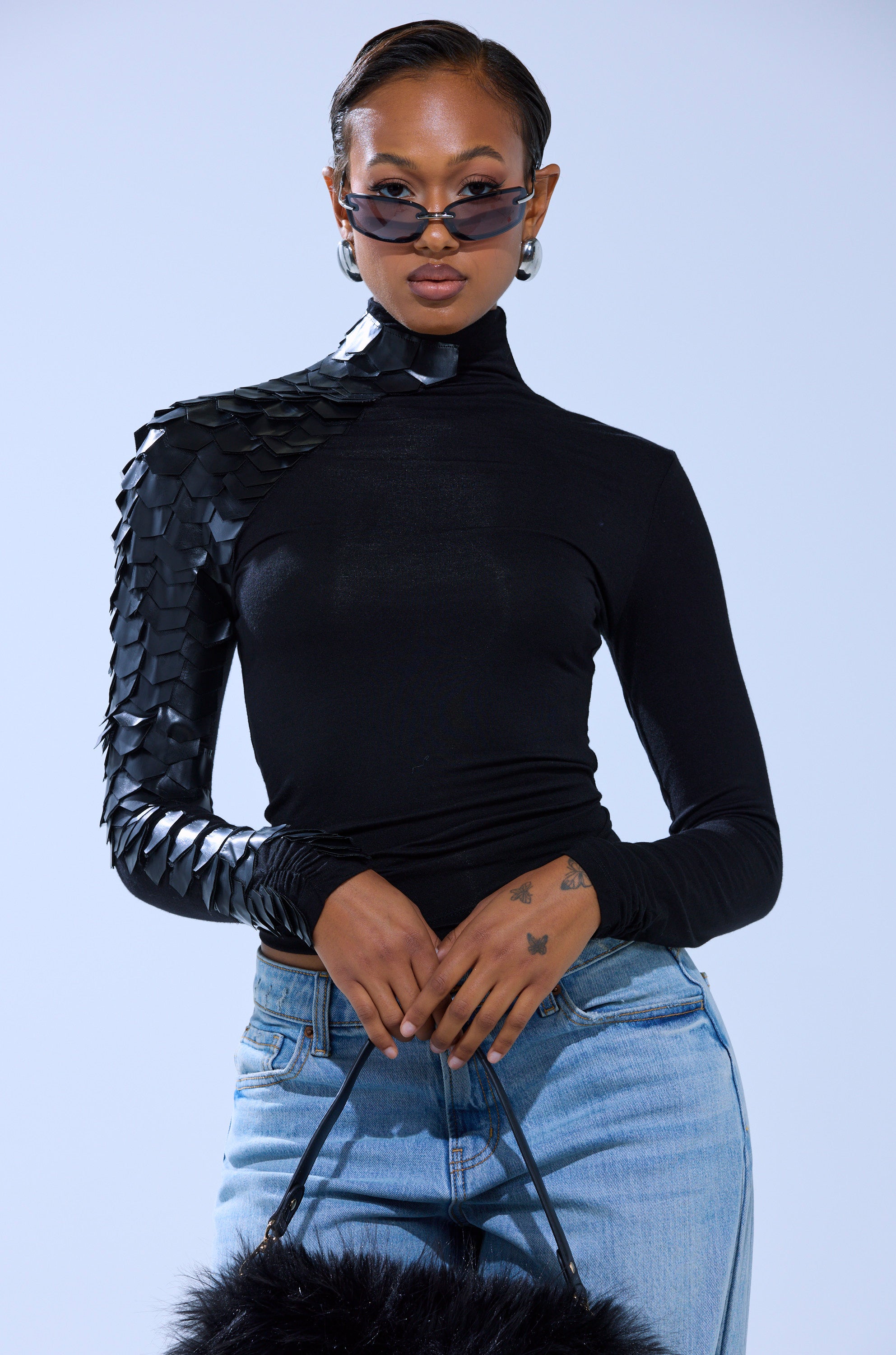 A person wears the NASTASIA SCALE TOP with geometric faux leather cutouts on one shoulder, styled with blue jeans, sunglasses, large hoop earrings, and a black furry bag against a light background.