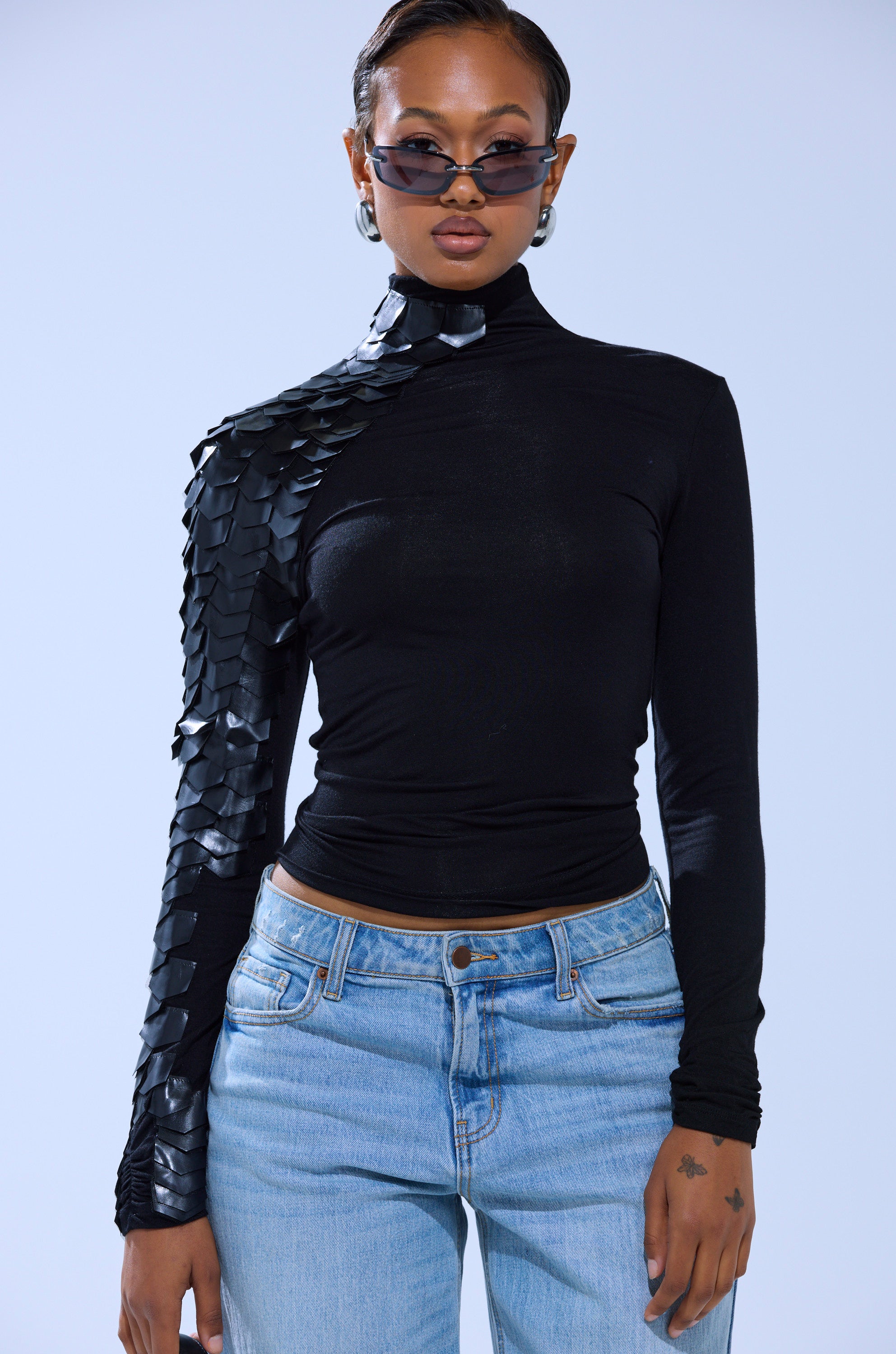 A woman in sunglasses, hoop earrings, the NASTASIA SCALE TOP with shiny textured details and geometric faux leather cutouts on one sleeve, and light blue jeans stands against a plain light background.