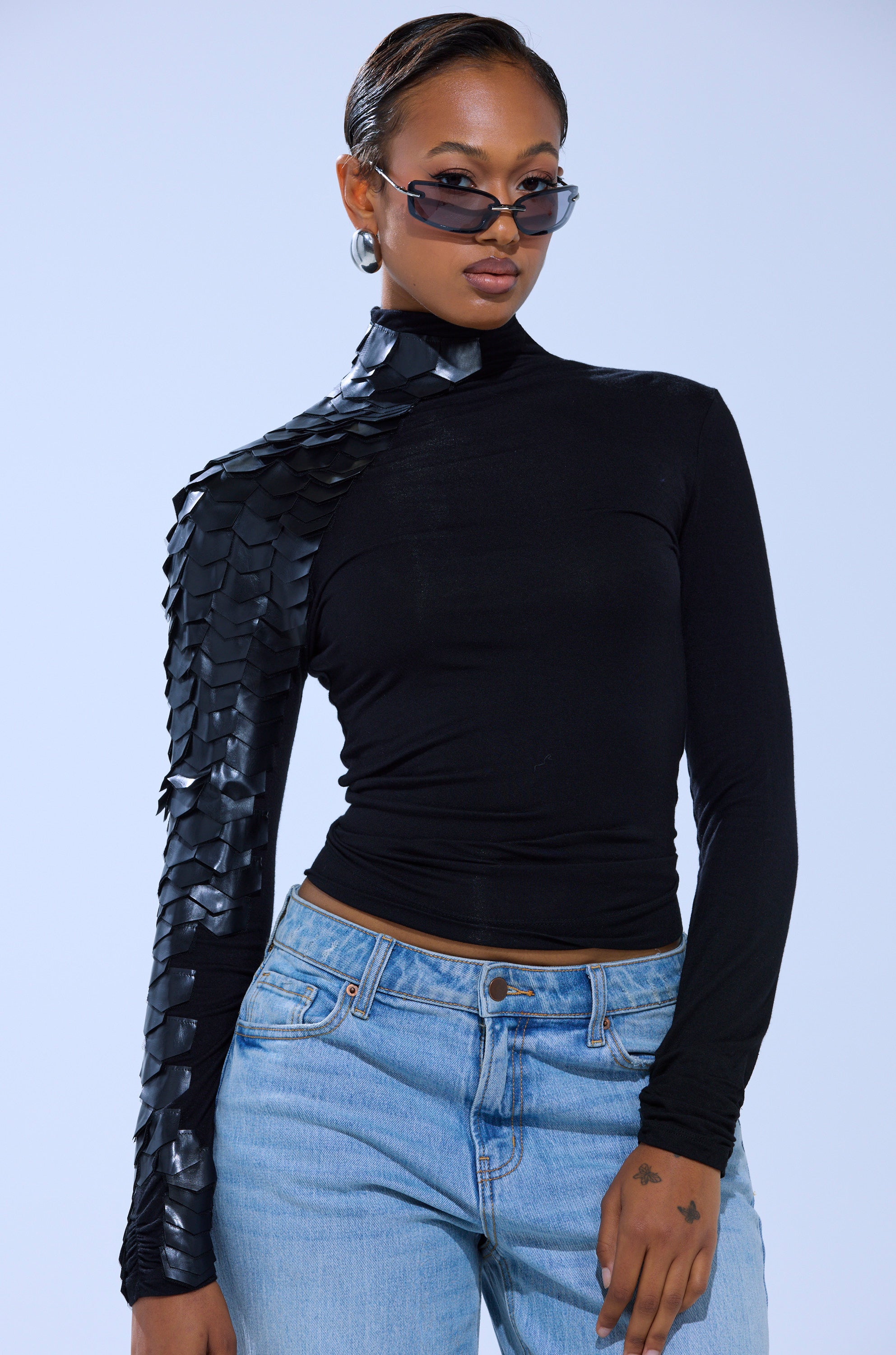 A person wearing sunglasses, a black NASTASIA SCALE TOP with shiny, textured scales on one shoulder and sleeve, and light blue jeans poses confidently against a plain light background.