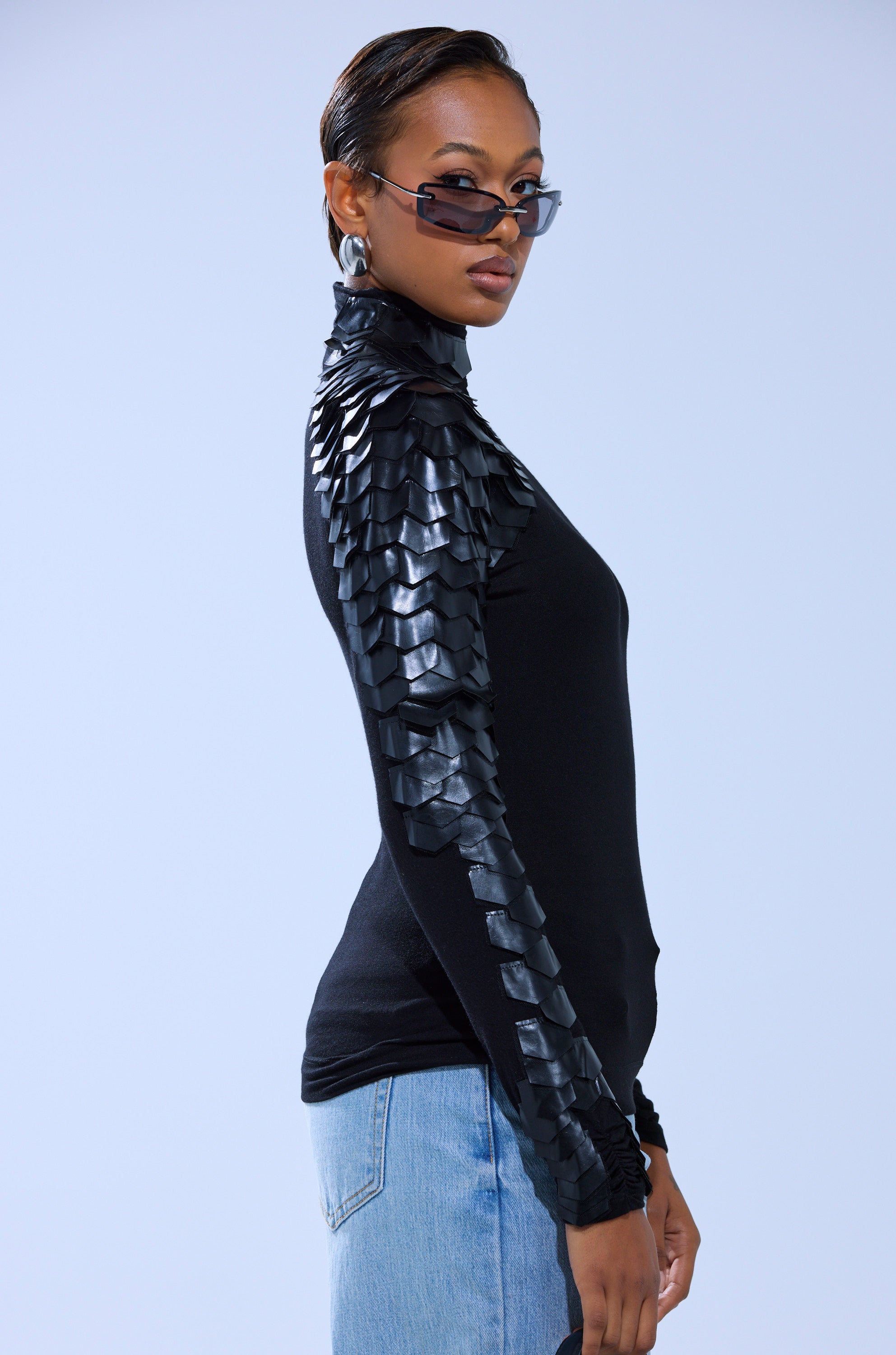 A woman in AKIRA Label sunglasses, the NASTASIA SCALE TOP with geometric faux leather cutouts, and light blue jeans stands against a plain background, looking confidently over her shoulder.