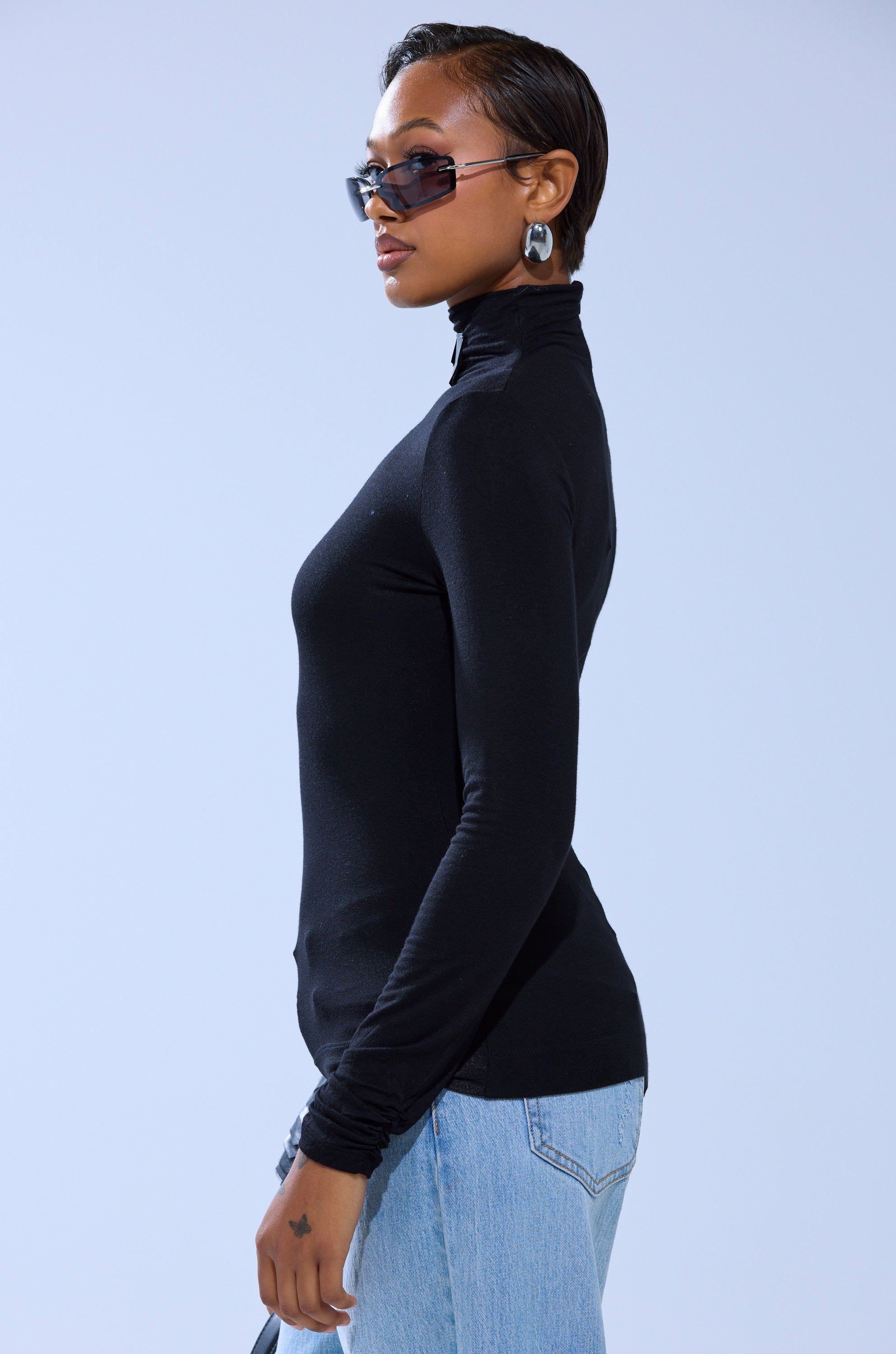 A person stands in profile against a plain light background, wearing sunglasses, silver hoop earrings, the AKIRA Label NASTASIA SCALE TOP in black, and light blue jeans.